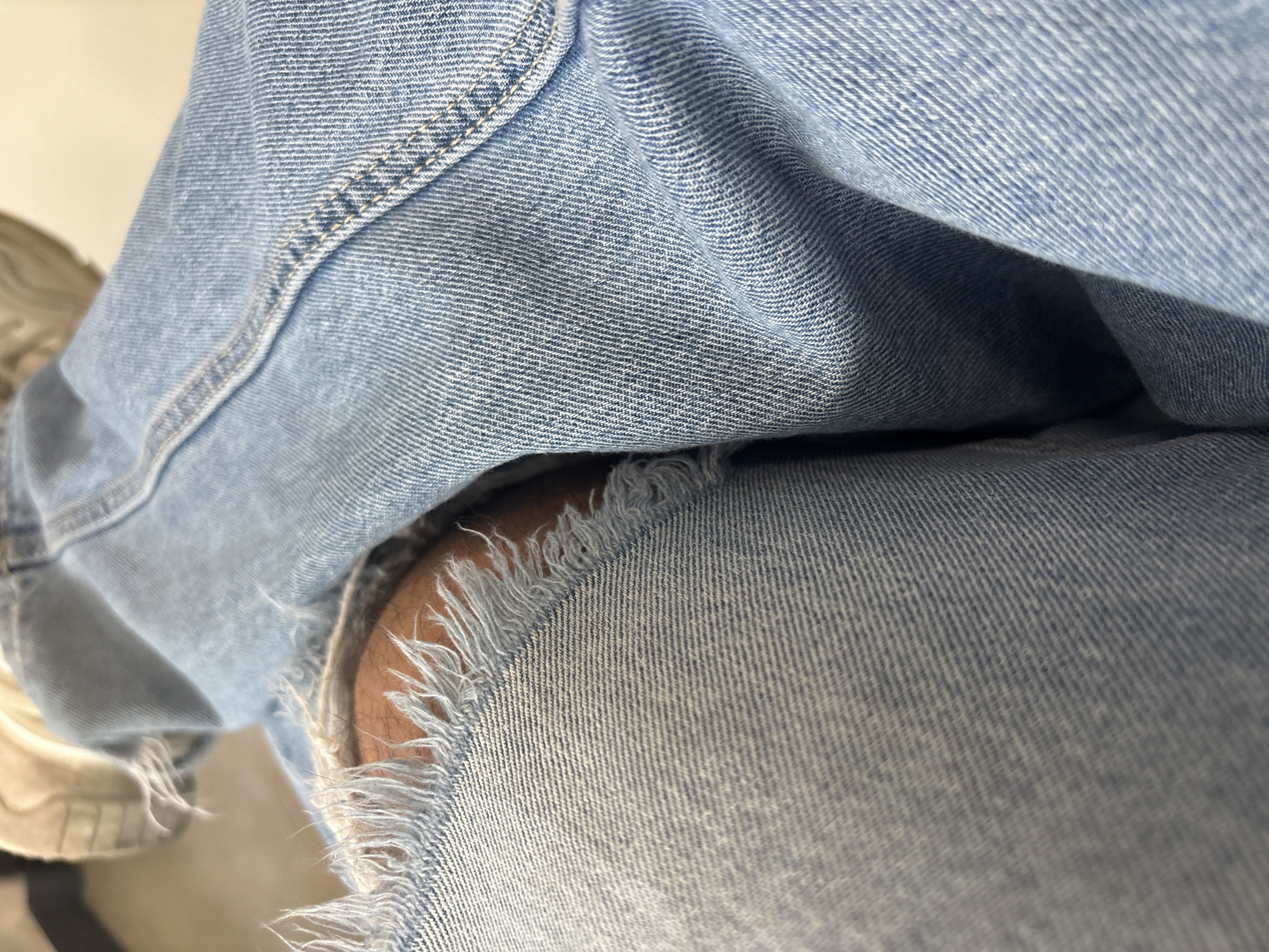 The image shows a close-up view of a person wearing light blue denim jeans with a ripped knee. The exposed skin of the knee is visible through the tear in the jeans. A small portion of a beige shoe is visible in the lower left corner of the image. No other details are discernible, and the location is not identifiable from the image.