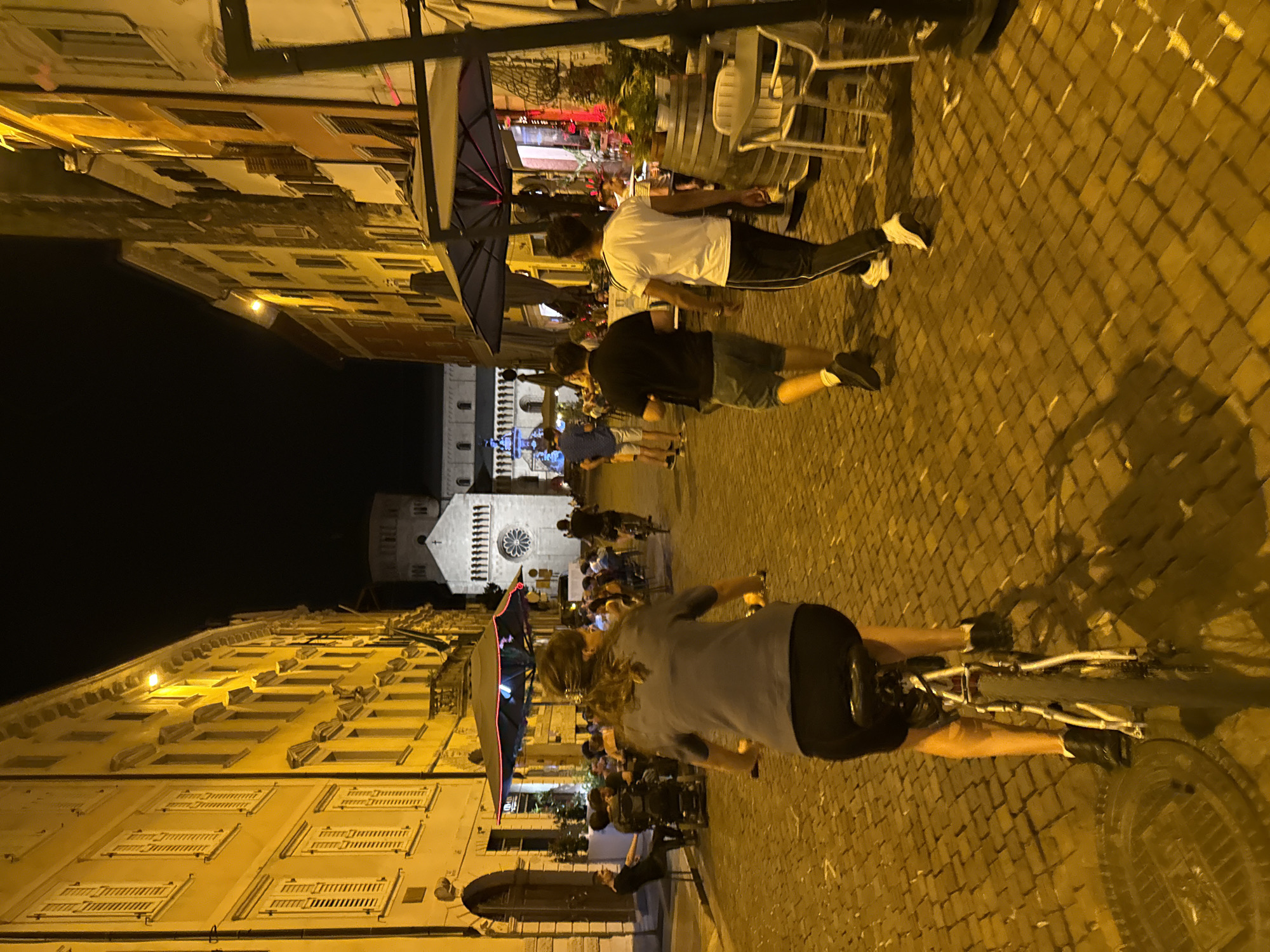The image depicts a street scene at night in Trento, Italy. The street is paved with cobblestones and lined with buildings on both sides, illuminated by warm yellow streetlights. On the left, a woman is riding a bicycle towards the viewer. She has dark hair pulled back and is wearing a grey top and black leggings. Her bicycle is silver and white.
In the background, several people are seated at tables outside a restaurant, enjoying drinks and conversation. Red and blue umbrellas are set up over the seating area. Further down the street, two men are walking away from the camera. One is wearing a black t-shirt and shorts, and the other is wearing a white t-shirt and black track pants.
In the distance, a large stone building with a domed tower is visible. This building appears to be a church or cathedral. The overall atmosphere is lively and social, suggesting a pleasant evening in the city.