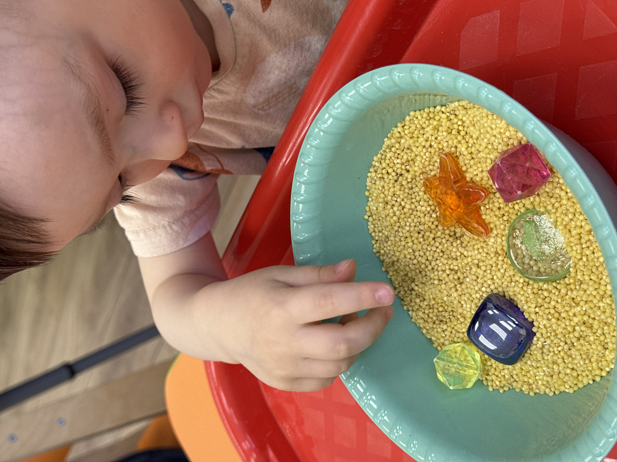 A close-up, high-angle shot captures a young child engaged in sensory play within an indoor setting, possibly a home or daycare in Izhevsk, Russia. The child's face, showing a closed eye with long eyelashes, nose, and cheek, is visible on the upper left, along with their left hand and arm. They are wearing a light-colored t-shirt with a subtle pattern.
The focal point of the image is a light blue (or teal) plastic bowl, featuring decorative scalloped edges, filled with a large quantity of small, light yellow granular material, likely millet or similar grain. Scattered throughout these grains are several colorful, translucent plastic or glass gems and figures. These include an orange starfish, a bright pink faceted gem, a clear or light green circular gem, a dark blue rectangular gem, and a light yellow faceted gem. The child's hand is reaching towards the contents of the bowl, indicating interaction with the sensory materials.
The bowl is placed on a vibrant red plastic surface, which appears to be a tray or part of a larger play unit. A portion of an orange surface is visible beneath the red on the lower left, and a wooden surface with a black strap is faintly discernible in the far left background. The scene is brightly lit, suggesting daytime. The child appears calm and focused, deeply engrossed in the exploratory activity. There is no visible text in the image.