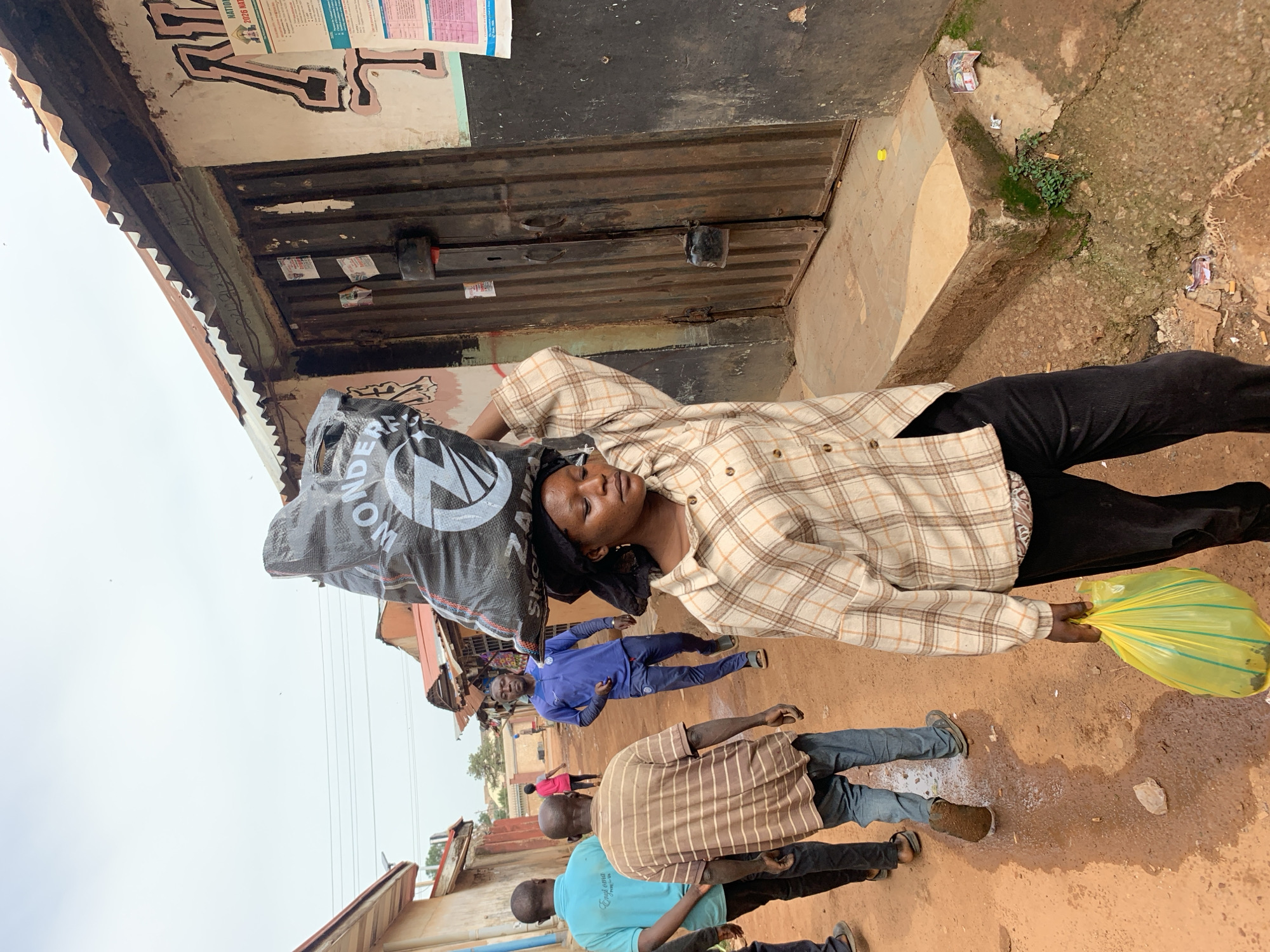 In Jos, Nigeria, a young woman walks down a dirt road, carrying a large black bag filled with goods on her head. She is dressed in a plaid shirt and black pants, and she holds a yellow plastic bag in her right hand. The road is lined with buildings, some of which appear to be shops or homes. The sky is overcast, and the overall atmosphere is one of a typical day in a bustling Nigerian city. The image captures a moment in time, showing the everyday life of people in Jos, Nigeria.