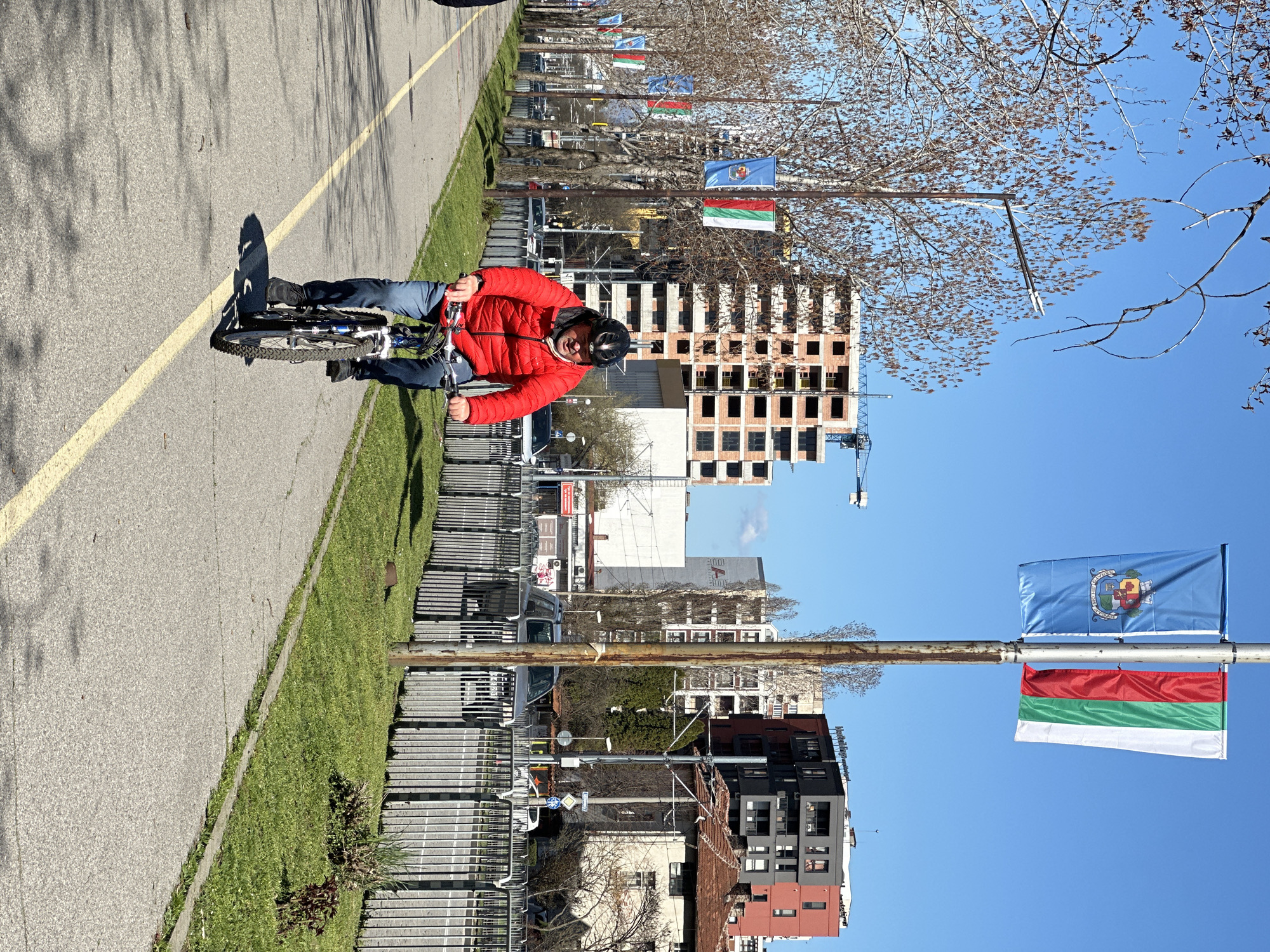 A person wearing a red jacket, blue jeans, and a helmet is riding a bicycle on a paved path. The path has a yellow line marking the edge. To the side of the path is a grassy area bordered by a metal fence. In the background are buildings, some under construction, and trees with bare branches. Flags of Sofia and Bulgaria are displayed on poles along the path. The image is taken in Sofia, Bulgaria.