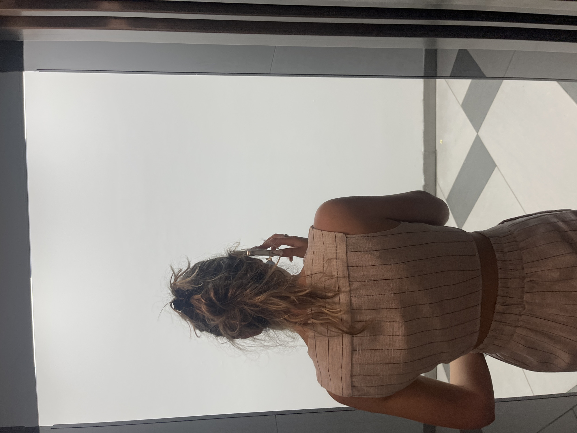 The image, rotated 90 degrees clockwise, shows a person viewed from behind, standing indoors and looking out through a large window or glass panel. The individual has light brown hair with blonde highlights, styled in a messy bun or updo. They are wearing a light, possibly beige or off-white, two-piece outfit. The sleeveless top features thin dark vertical stripes, while the bottom piece, which appears to be a skirt or shorts, has thin dark horizontal stripes, with a visible midriff section. The person's left hand is raised towards their head, appearing to hold a hair clip or a similar small item, while their right arm is partly visible.
Beyond the window, the environment is predominantly bright white, suggesting either an intensely lit outdoor sky, perhaps on an overcast day, or a highly reflective surface. To the right, a clear black and white checkered floor pattern is visible through the glass. The framing of the window or door is dark grey or black with some lighter metallic accents. The overall scene suggests a modern interior looking out onto another area. No visible text is present in the image. This scene is set in Blagoevgrad, Bulgaria.
