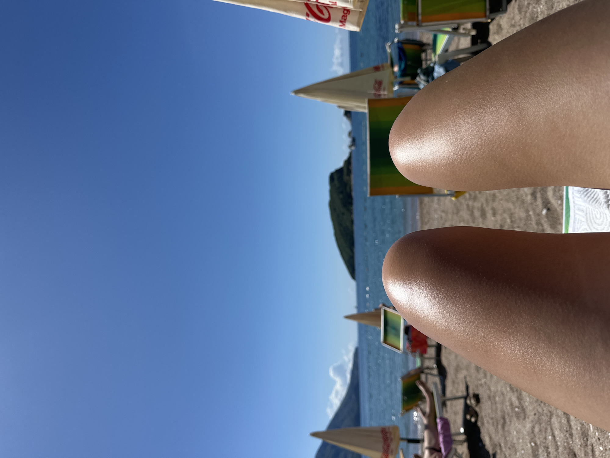 The image depicts a sunny beach scene, likely in Budva, Montenegro. The focus is on two legs belonging to a person relaxing on the beach. In the background, there is a clear blue sky, the sea, and a small island or landmass. Several beach umbrellas and lounge chairs are visible, suggesting a tourist area. Some people are present on the beach. The sand appears to be light brown.