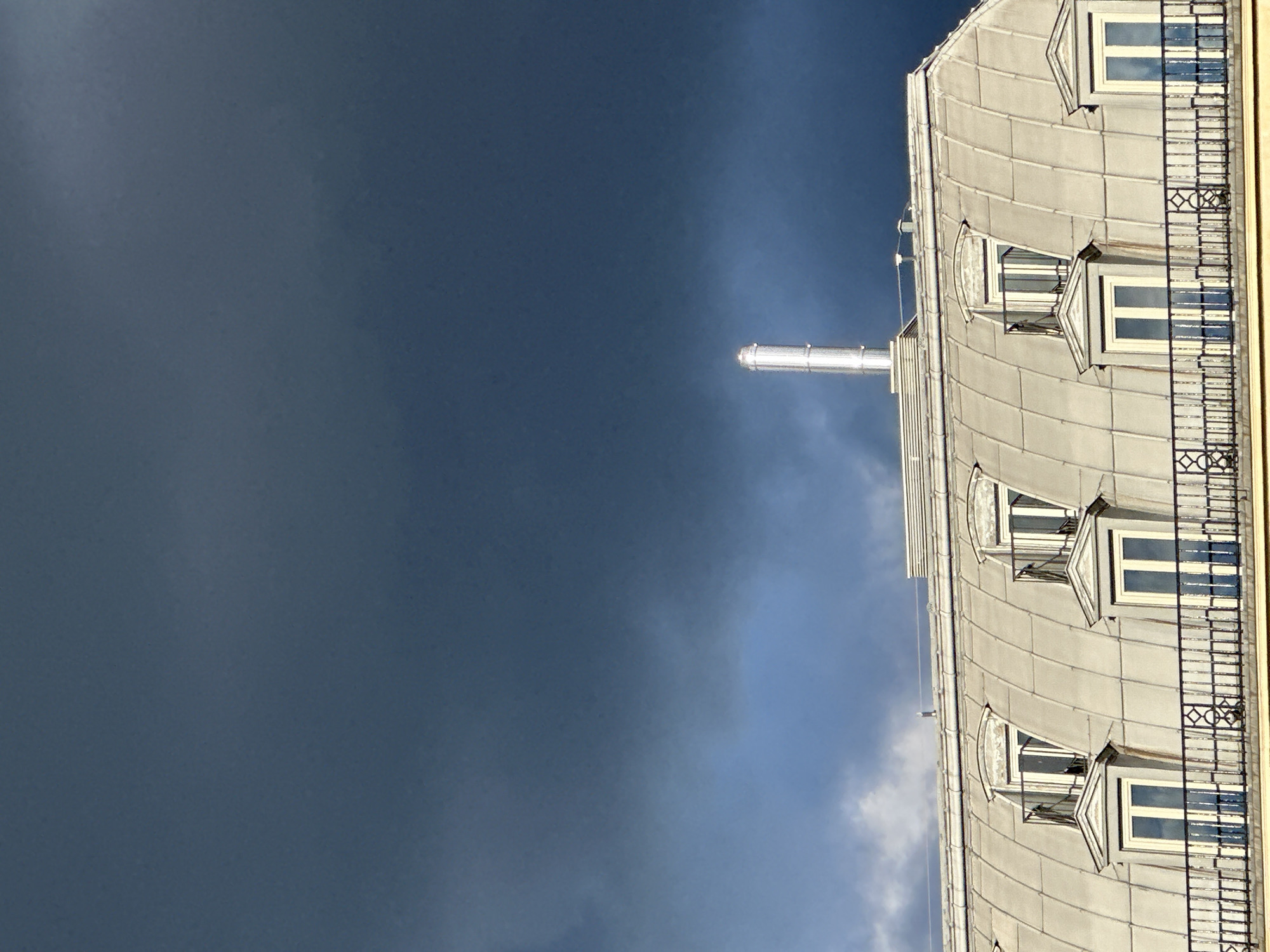 The image shows the roof of a Parisian building with a curved, metal-tiled roof. Four windows are visible, each with a small, triangular projection above. A wrought-iron balcony runs along the side of the building. A metal chimney protrudes from the roof. The sky is a dark, stormy blue on the left, transitioning to a lighter blue on the right, suggesting an approaching storm. The location is likely in Paris, France, based on the architectural style.