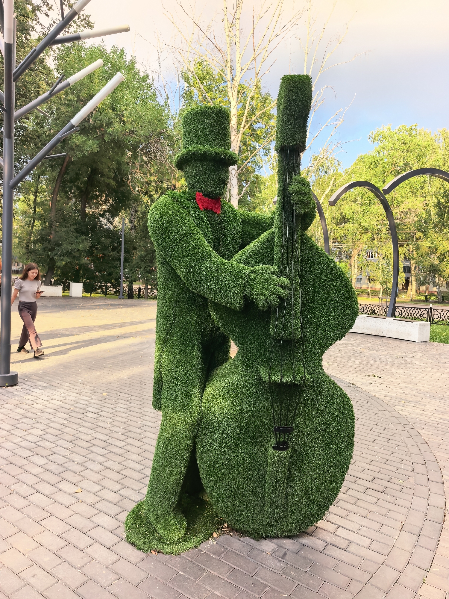 An outdoor shot captures a topiary sculpture of a person playing a double bass. The sculpture is made of green grass and depicts a figure wearing a top hat and a red bowtie, with their arms positioned as if playing the instrument. The double bass is also rendered in topiary form. The sculpture stands on a paved walkway made of brick-like stones. To the left of the sculpture, a young woman with dark hair, wearing a grey top and dark trousers, walks away from the camera, looking at her phone. Behind the sculpture, trees and foliage create a park-like setting. In the background, there are decorative metal arches and more trees. The sky above is partly cloudy with warm sunlight. The sculpture is located in the city of Sterlitamak, Russia.