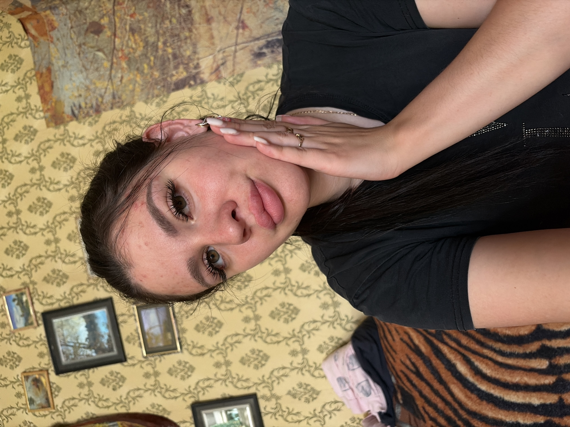The image shows a close-up selfie of a young woman with dark hair and fair skin. She is wearing a black t-shirt and has her right hand on her cheek. She is wearing earrings and rings on her fingers. The background appears to be a wall with floral wallpaper and several small framed pictures. There is also a glimpse of a tiger-striped patterned cushion or blanket in the lower left corner of the image. The lighting suggests an indoor setting, possibly a room in a home. There is no visible indication of the city of Большой Букор, Russia, or any specific location.