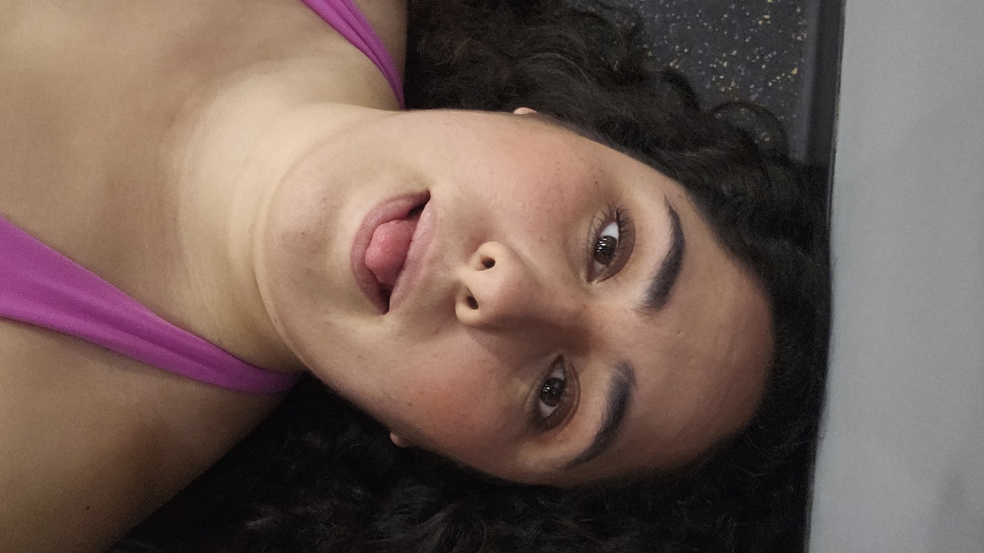 The image depicts a close-up, vertically oriented shot of a single individual, a young adult female with an olive skin tone, dark brown eyes, and dark, curly hair. Her head is tilted to the viewer's right. She is seen extending her tongue from her mouth, with her gaze directed towards the viewer. She wears a fuchsia-colored strap garment, consistent with a sports bra or tank top, visible across her upper chest and shoulder. Distinct tan lines are present on her décolletage. The background on the upper left side of the frame is a dark, speckled surface, likely a rubberized gym floor with yellow and white flecks. The background on the lower right side is a plain, light gray wall or partition. The scene is indoors, consistent with a fitness facility. The image does not contain identifiable visual markers to specify its location within the city of Puebla, Mexico.