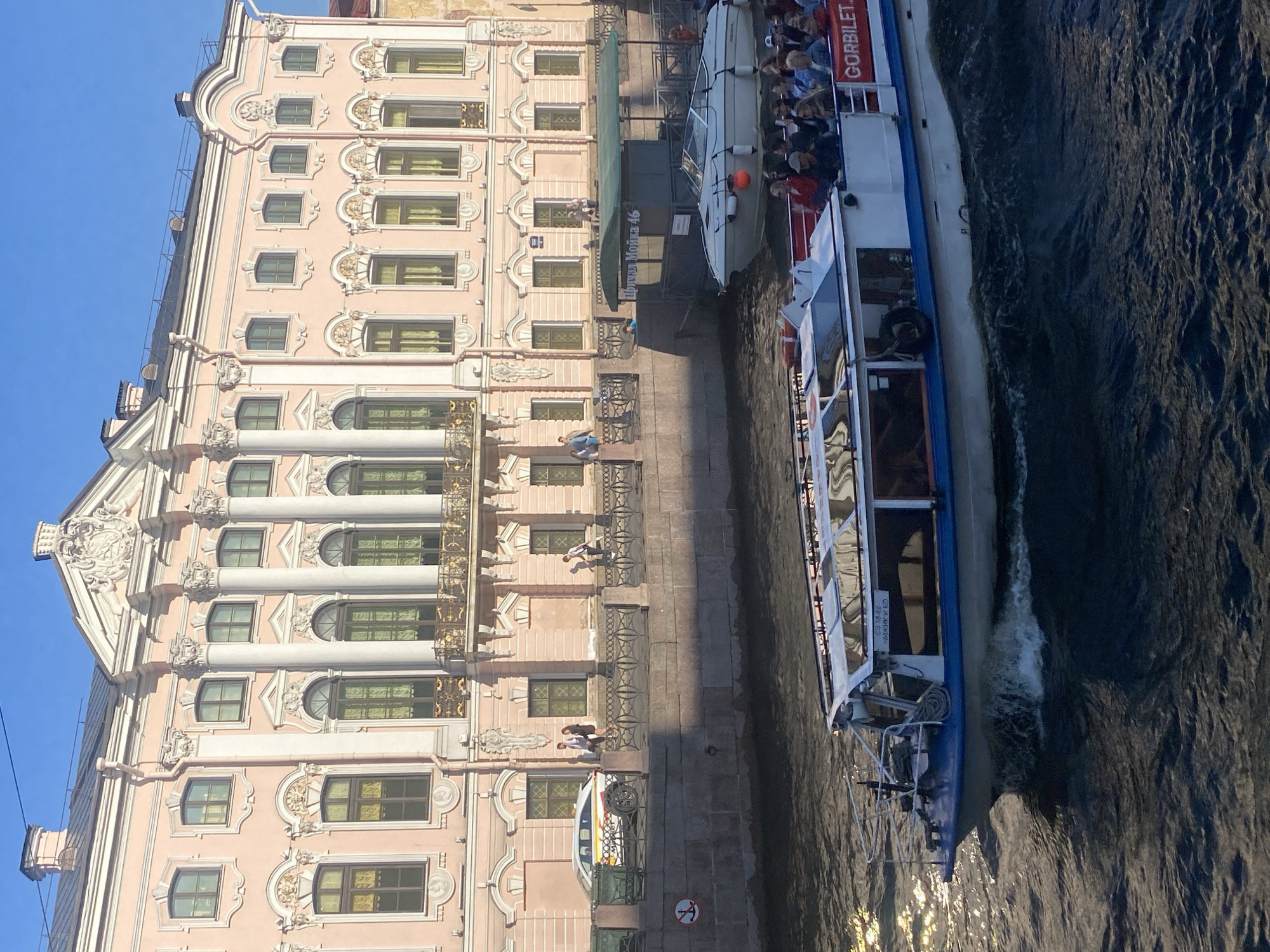 The image depicts a sunny day scene on a canal in Saint Petersburg, Russia. In the foreground, a white and blue tour boat with passengers is moving through the water, creating a wake. The boat has a covered section with windows and an open-air area at the back. The word "GORBILET" is visible on the side of the boat.
In the middle ground, a pale pink building with classical architectural elements, including columns, elaborate window frames, and decorative moldings, lines the canal. A few people are walking along the embankment in front of the building. A white car with yellow and red stripes is parked on the embankment.
Further back, another smaller white boat is docked near a green awning with Russian text that appears to be "Причал Мойка 46" (Pier Moika 46). The canal water is dark and rippled, reflecting the sunlight. The sky is clear and blue.