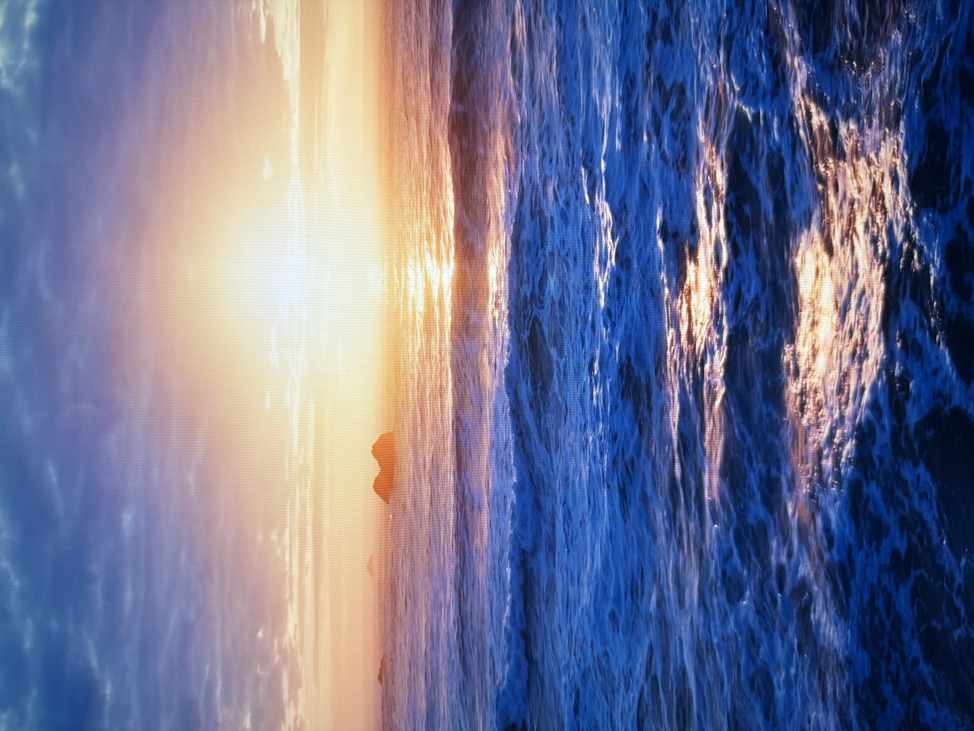 The image depicts a serene ocean sunset with a vibrant sky transitioning from deep blue to hues of orange and yellow as the sun dips below the horizon. The ocean surface is a deep blue, reflecting the warm tones of the sunset, with gentle waves creating white foam. In the distance, silhouetted rock formations rise from the water. There is no indication or depiction of the city of Kyiv, Ukraine, in this image.