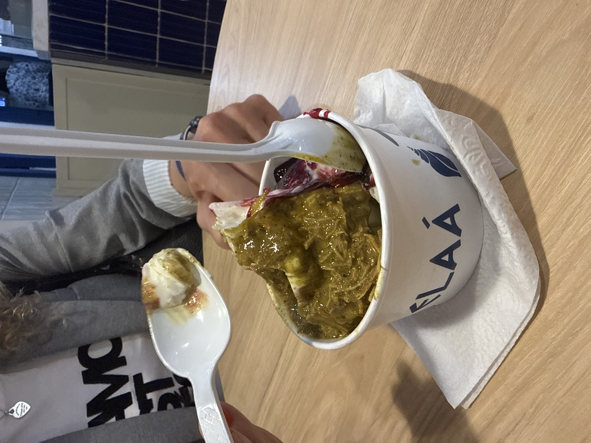 A person is eating ice cream from a white cup labeled "ELAA," likely in Puebla, Mexico, though the image itself offers no direct location confirmation beyond the brand which has locations in Puebla. The ice cream appears to have a pistachio base, with red sauce or fruit, and noodles. A second spoon scoops out the ice cream. The scene is set on a wooden table with a white napkin nearby. The person wearing a gray sweater with a white collar and visible part of a T-shirt, and a watch is partially visible. The cup of ice cream is the central focal point. A tiled wall is located in the background.