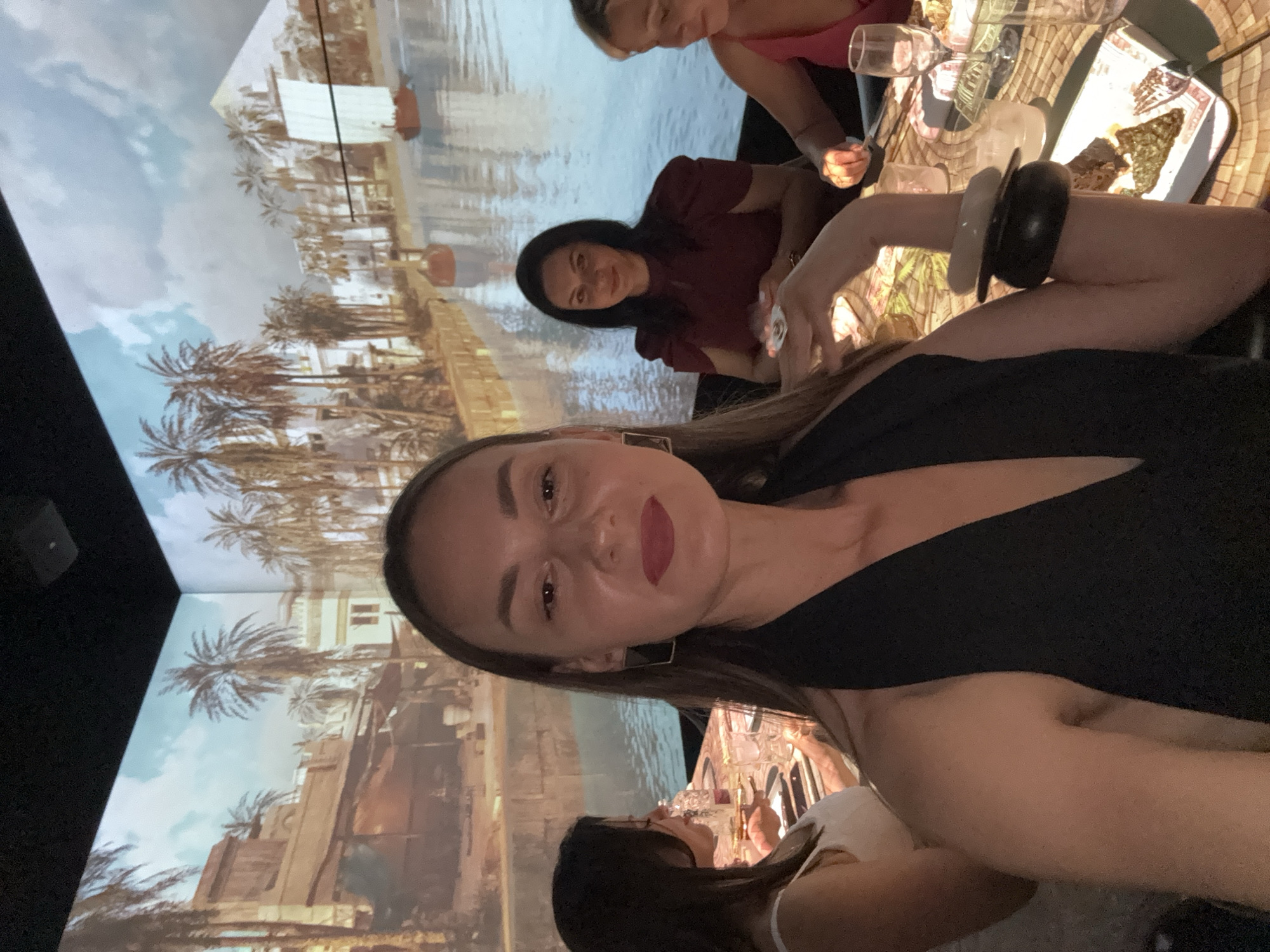 The image depicts a group of women seated at a table in what appears to be a restaurant or event space. The background is a large projection of an ancient Egyptian-themed scene, featuring palm trees, buildings, a river, and a pyramid.
In the foreground, a woman with long brown hair, wearing a black halter top and large rectangular earrings, is taking a selfie. She has dark lipstick on. To her left, another woman with dark hair, wearing a white top, is partially visible, looking down at the table.
Behind the selfie-taker, to the right, a woman with dark, shoulder-length hair is wearing a maroon dress and smiling. She is holding a small white object in her hands. To her right, another woman with blonde hair, wearing a pink top, is visible, also looking towards the table.
The table is set with plates, cutlery, glasses, and what appears to be food. The woman in the foreground is wearing two stacked bracelets on her left wrist, one black and one light-colored. The overall lighting suggests an indoor setting with ambient light and the projected background illumination. The projection covers the walls and possibly the ceiling, creating an immersive environment.