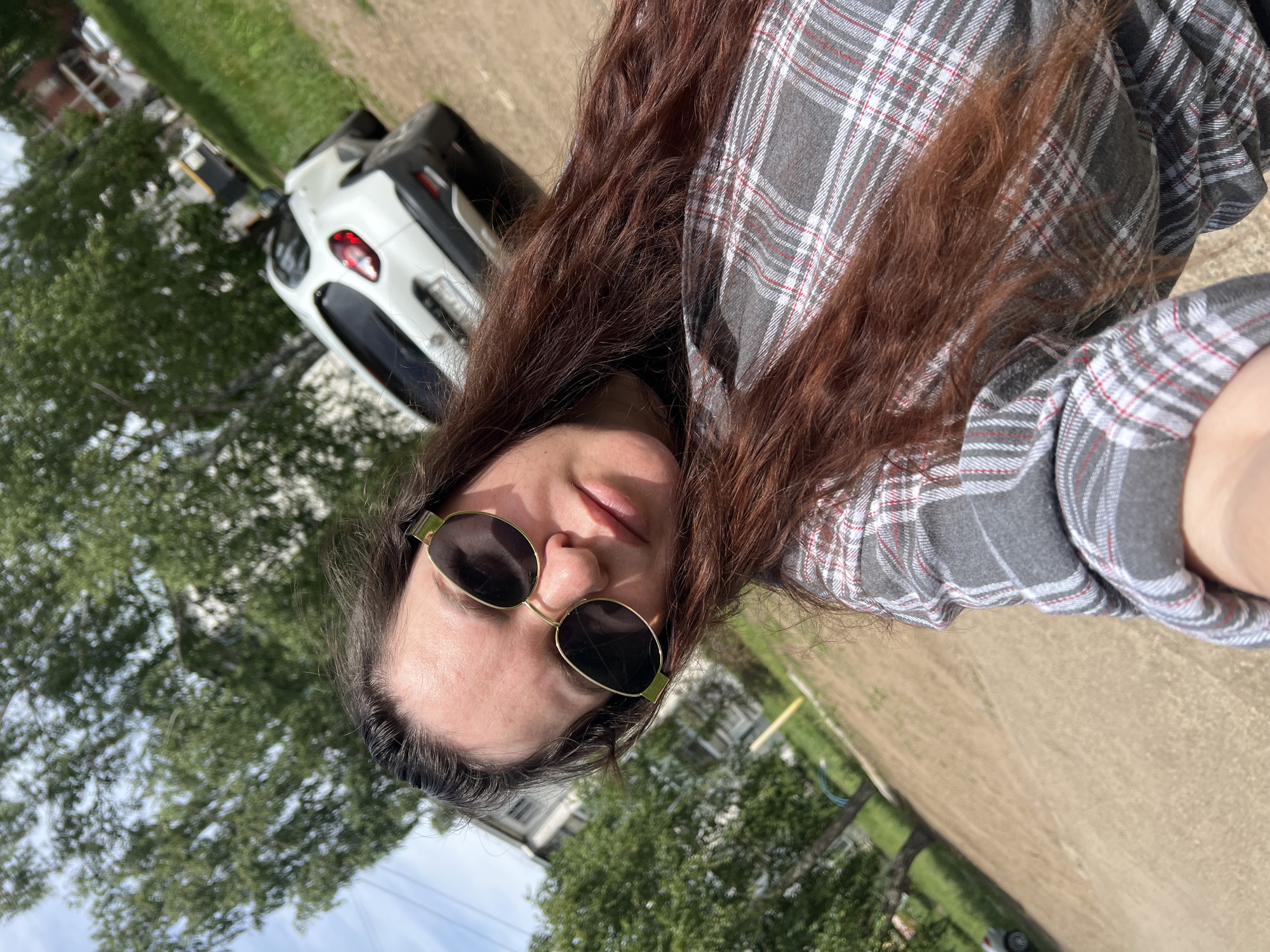 This brightly lit, angled selfie features a person with long, wavy dark brown hair, wearing oval-shaped sunglasses with gold frames and a grey plaid button-up shirt with thin red and white lines. The person's head is tilted, and their face is partially obscured by the sunglasses. Sunlight casts shadows on their face, indicating a sunny outdoor setting.
In the background, a white compact car or SUV is partially visible, parked on a grassy or unpaved area. Lush green trees are prominent throughout the scene, with some closer to the foreground and others in the midground and distance. The immediate foreground and midground consist of a light brown, unpaved dirt or gravel path. In the far background, behind the trees, indistinct residential buildings can be seen.
The scene depicts an outdoor environment, likely a rural or suburban area in Noviy, Russia, characterized by natural elements like trees and unpaved ground, alongside distant structures. The clear skies and strong sunlight confirm it is daytime. No specific activity beyond taking a selfie is apparent, and the person's expression is neutral. There is no visible text in the image.