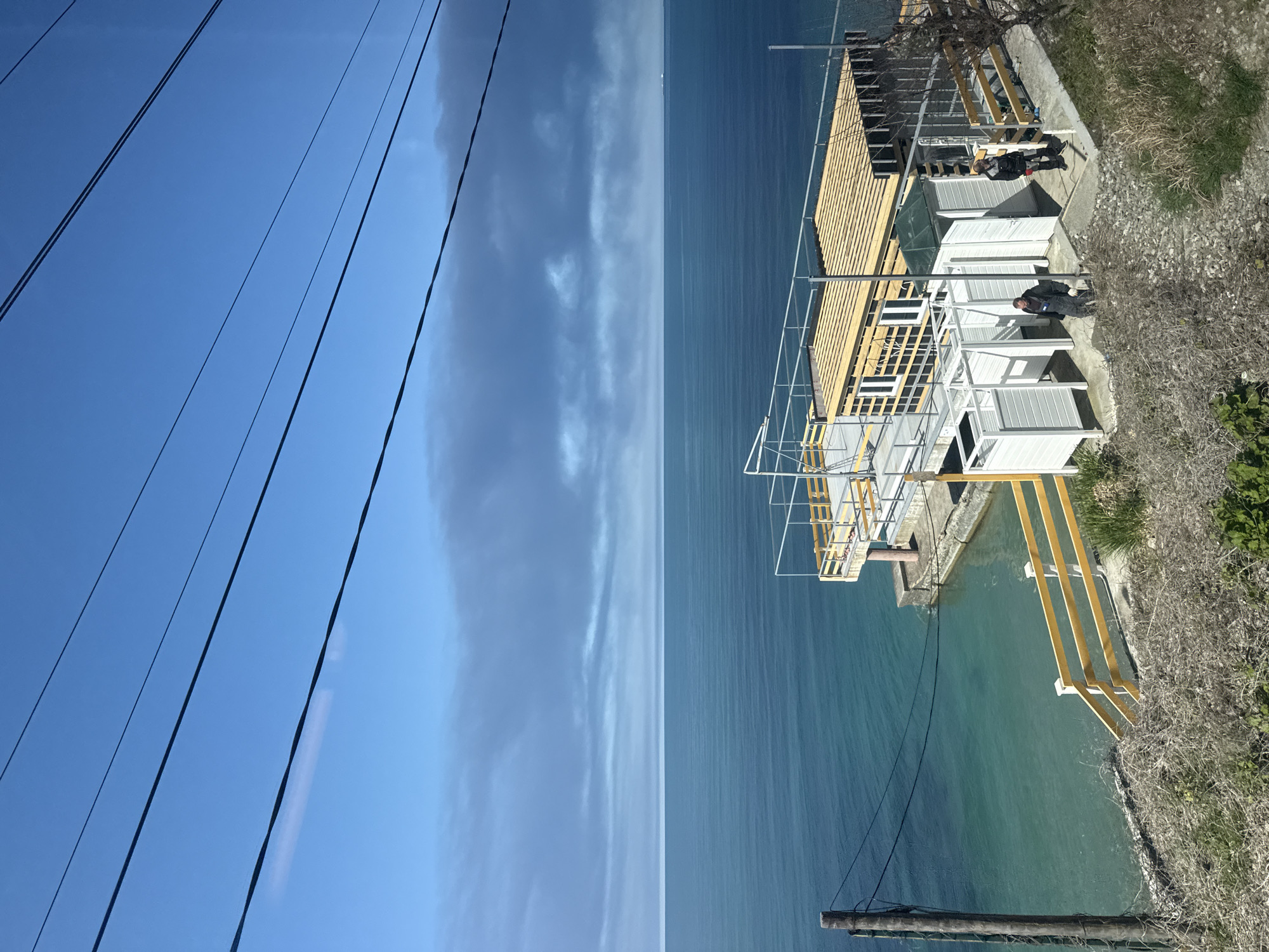 This image shows an elevated view of a seaside structure in an unknown city, unknown. The structure consists of white and yellow supports and enclosures, with some wooden framework on top. Two people are present on the structure. The body of water next to the structure has a dark to light blue color gradient. The sky in the image is divided by water, with the left side being a clear blue with power lines, and the other side containing a sky with clouds. Vegetation and concrete can be seen on the right of the image. In the lower right, there is a utility pole.