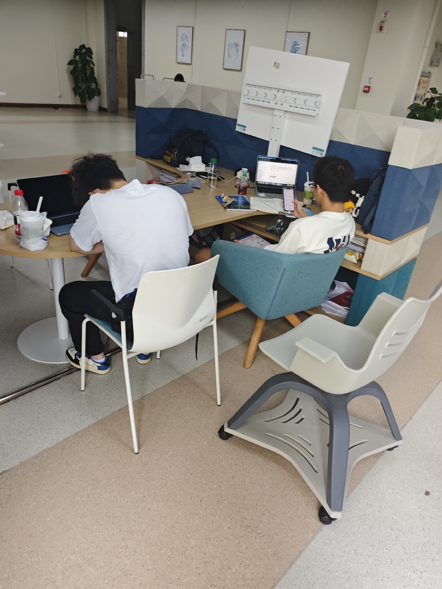 Two individuals are seated at a desk in what appears to be a modern office or study space. The person on the left, wearing a white t-shirt and black pants, is hunched over a laptop, seemingly asleep or resting their head on their arm. They are seated on a white plastic chair. On the desk in front of them are a laptop, a bottle of water, a cup, and some papers. The person on the right, wearing a white long-sleeved shirt, is seated in a blue armchair and is looking at their mobile phone. They are positioned in front of a large white screen mounted on a stand. The desk area is sectioned off with blue and white partition panels. In the foreground, a gray and white office chair with wheels is visible. The floor has a speckled pattern of light brown and gray tiles. The environment suggests a shared workspace or a study lounge. The image location is confirmed as the Chongqing High-tech Zone, China.