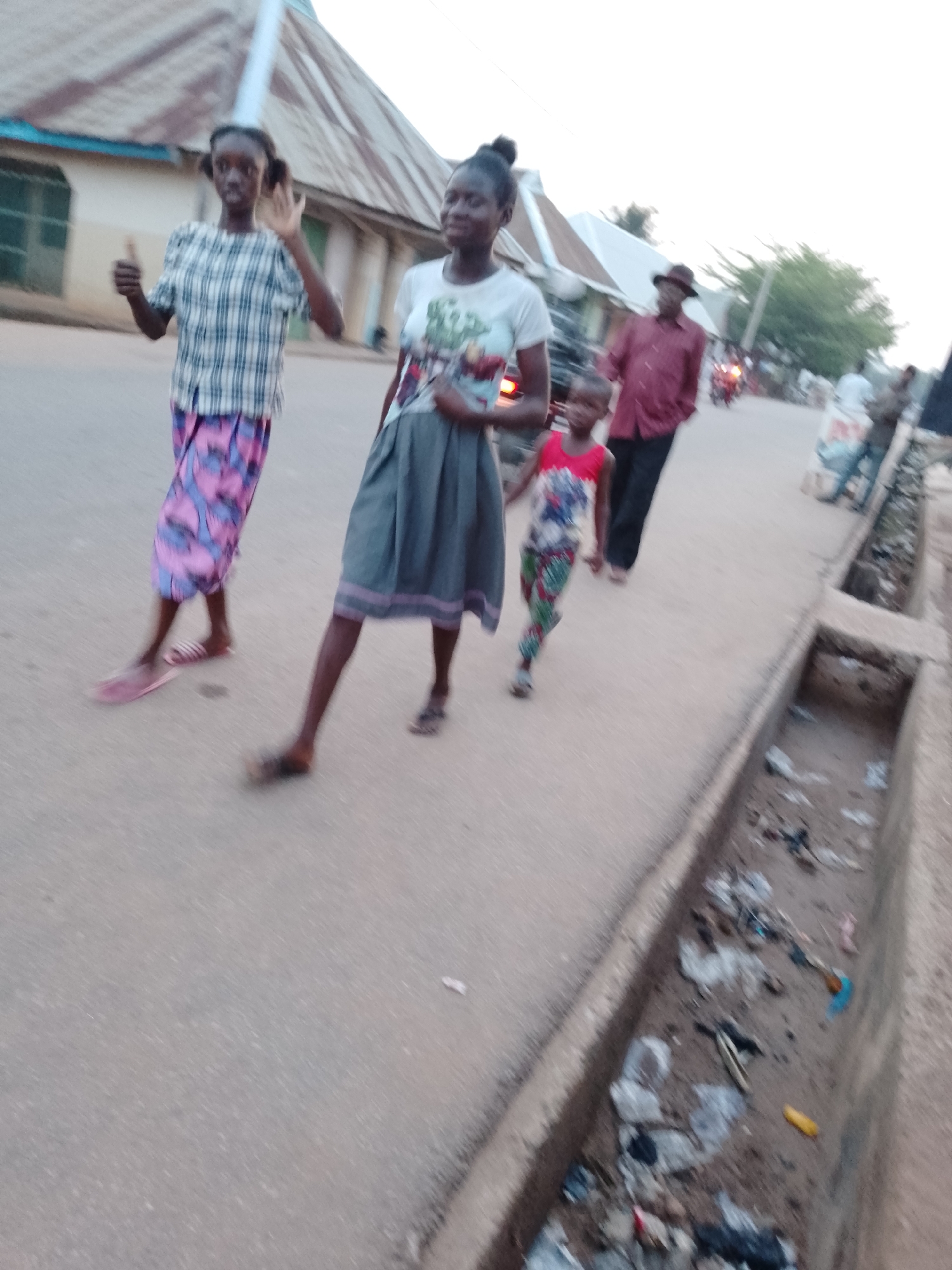 The image shows a scene on a street in Akwanga, Nigeria. Four individuals are walking along the paved street. In the foreground, a young woman wearing a plaid shirt and a patterned pink and blue skirt is giving a thumbs-up gesture. Beside her, another young woman with her hair in a bun is wearing a white T-shirt and a grey skirt. A young child, wearing a patterned red top and patterned green pants, is walking alongside the second woman. Behind them, a man wearing a hat and a red shirt is walking. A concrete drainage ditch filled with trash runs along the right edge of the street. Buildings with metal roofs are visible in the background, along with other people and vehicles.