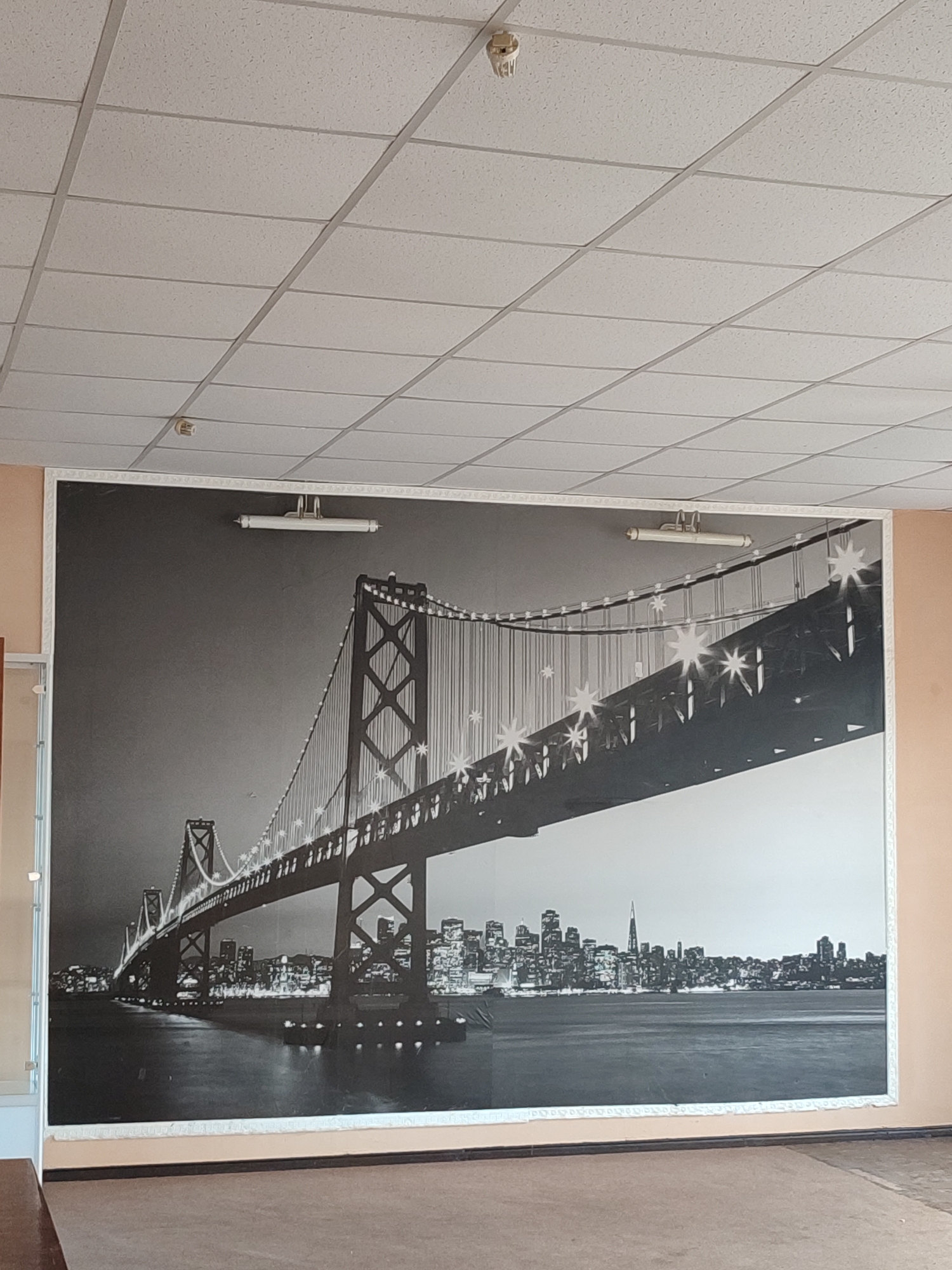 This media file shows a large mural of the Golden Gate Bridge at night, spanning an entire wall. The mural is a black and white photograph with bright lights illuminating the bridge and the city skyline in the background, suggesting a nighttime scene. Two fluorescent light fixtures are mounted on the wall above the mural. The ceiling is a typical drop ceiling with acoustic tiles and a smoke detector. The floor in front of the mural appears to be carpeted. The surrounding walls are painted a light peach color, and a portion of a white door frame is visible on the left side of the image. The overall setting appears to be an indoor space, possibly a hallway or a common area in a building. There is no visible text in the media file. The image appears to be taken in Shyshaky, Ukraine, but this is not evident from the visual content itself.