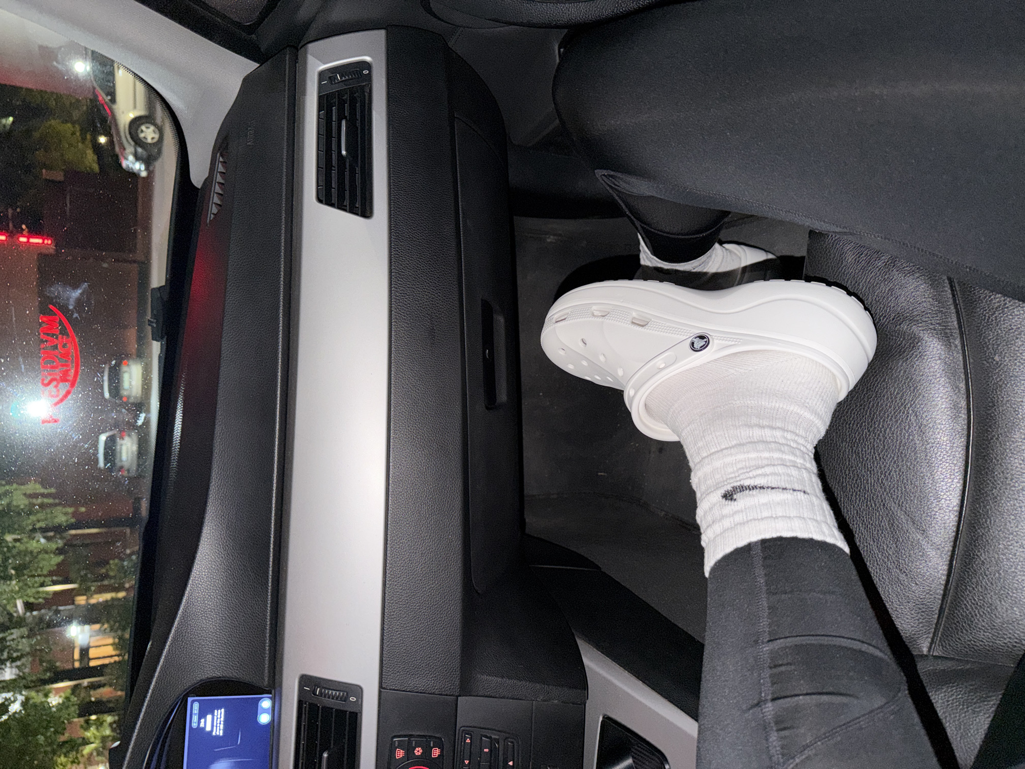 The image shows the interior of a car, specifically the passenger side. A leg wearing black leggings, a white Nike sock, and a white Croc shoe is visible on the seat. The car's dashboard and interior panels are black and silver. Through the car's windshield, a blurry view of the outside is seen, including trees, cars, and a building with a sign that reads "SIDDIQ PLACE." The location is not in Lovech, Bulgaria.