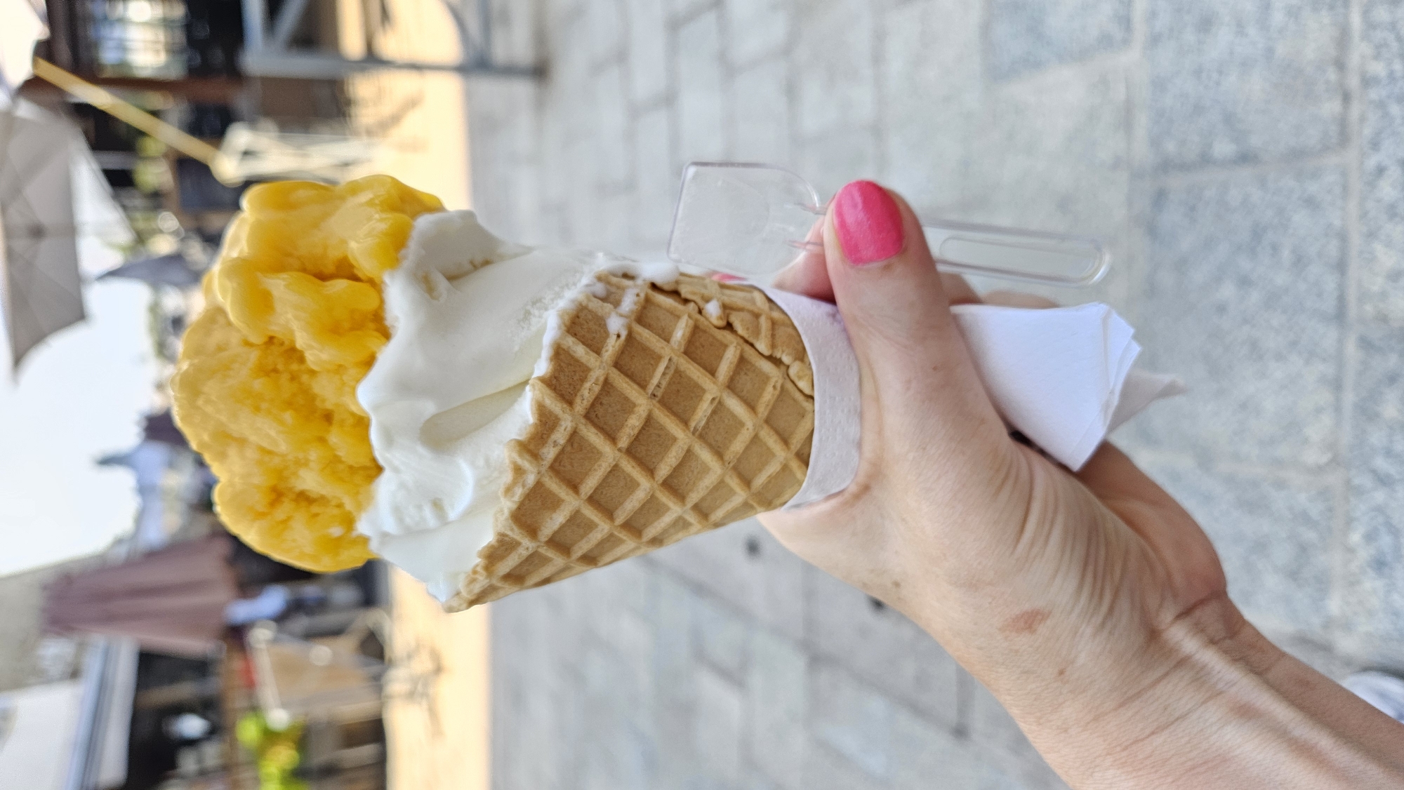 A hand with bright pink fingernails holds a waffle cone filled with two scoops of ice cream, one white and one yellow. The ice cream appears to be melting slightly. A small plastic spoon is held between the thumb and index finger, attached to a white napkin wrapped around the base of the cone. The background is blurred, suggesting an outdoor setting with paved ground and structures typical of a town or city. The scene is likely captured in the city of L'Île-Rousse, France, given the context.