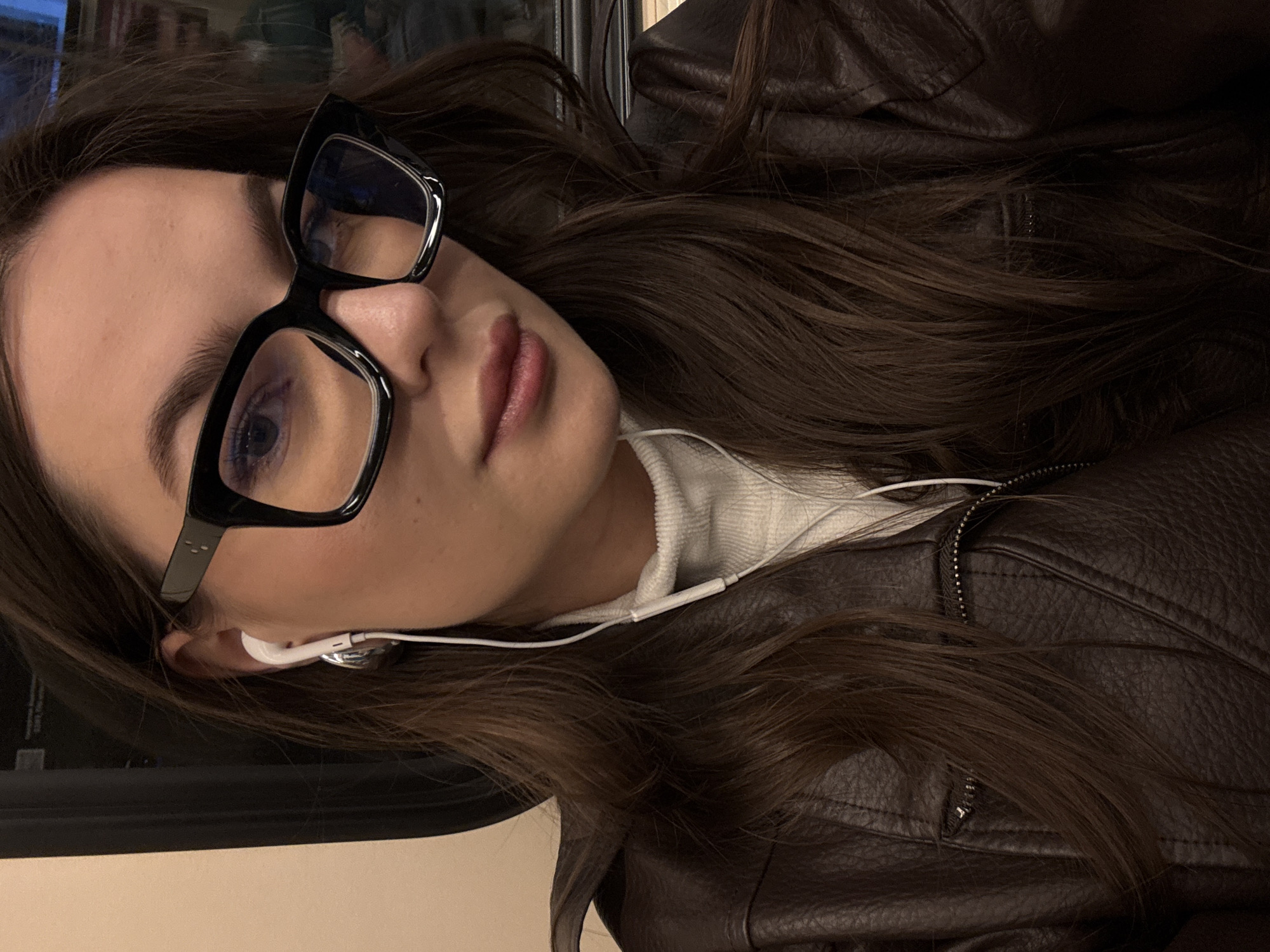 A young woman with long brown hair and black-framed glasses is on what appears to be a public transport vehicle. She is wearing a white turtleneck sweater and a brown leather jacket. She has white earbuds in her ears. The background suggests a moving vehicle with a window. There is no information that identifies the location. Therefore, it's not possible to conclude the photo was taken in Moscow, Russia.