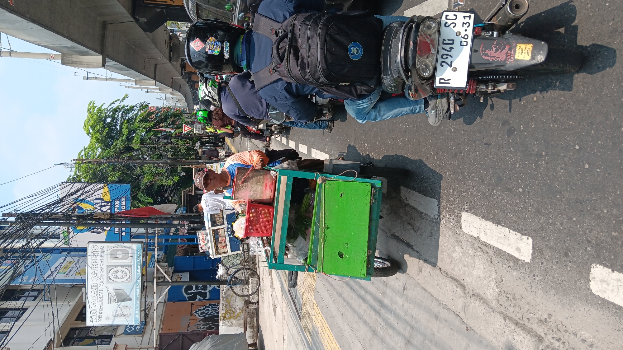 A street vendor pushing a green cart is seen in the middle of traffic on a sunny day in South Jakarta, Indonesia. The cart is laden with various items, including red baskets, a large plastic container, and what appear to be produce or packaged goods. The vendor, a man wearing a brown cap and an orange and blue shirt, is looking towards the camera. Behind him, several motorcycles are also in traffic. One motorcycle, closest to the viewer, has a rider wearing a blue jacket and jeans, with a black backpack. The motorcycle has a license plate that reads "R 294G SC 06.31" and features stickers on its rear fender. In the background, buildings with various signs are visible, including one for "MOLADAD CARPETS". Overhead, a concrete overpass and numerous electrical wires dominate the skyline. The road is asphalt with visible white lane markings.