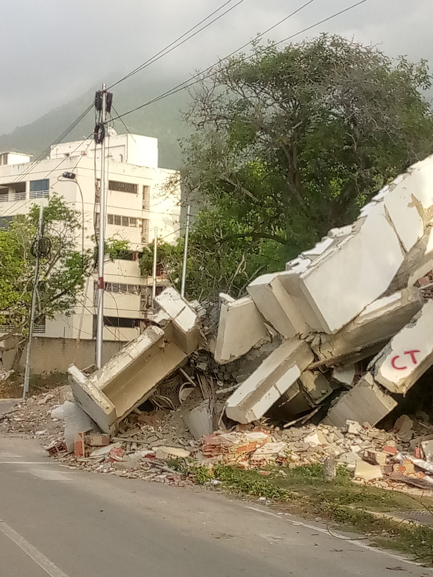 The image depicts the aftermath of a significant natural disaster, likely an earthquake or landslide, in Caraballeda, Venezuela. In the foreground, a pile of debris from a collapsed concrete structure dominates the scene. Large concrete slabs, some bearing red markings that appear to be "CT," are jumbled together. Beneath these slabs are smaller pieces of concrete, bricks, and twisted rebar, suggesting extensive structural failure. A section of asphalt road is visible in the lower part of the frame, with a white line marking the edge.
Behind the debris, a multi-story building stands partially intact but shows signs of damage, with visible cracks and missing sections. Trees with green foliage are scattered throughout the background, some appearing to have been uprooted or damaged. Power lines and utility poles are also visible, some leaning or damaged. In the far background, a hazy mountain range can be seen, shrouded in what appears to be a light fog or dust. The sky is overcast, contributing to the somber atmosphere of the scene. There are no people visible in the image. The overall impression is one of devastation and destruction, characteristic of a major catastrophic event.