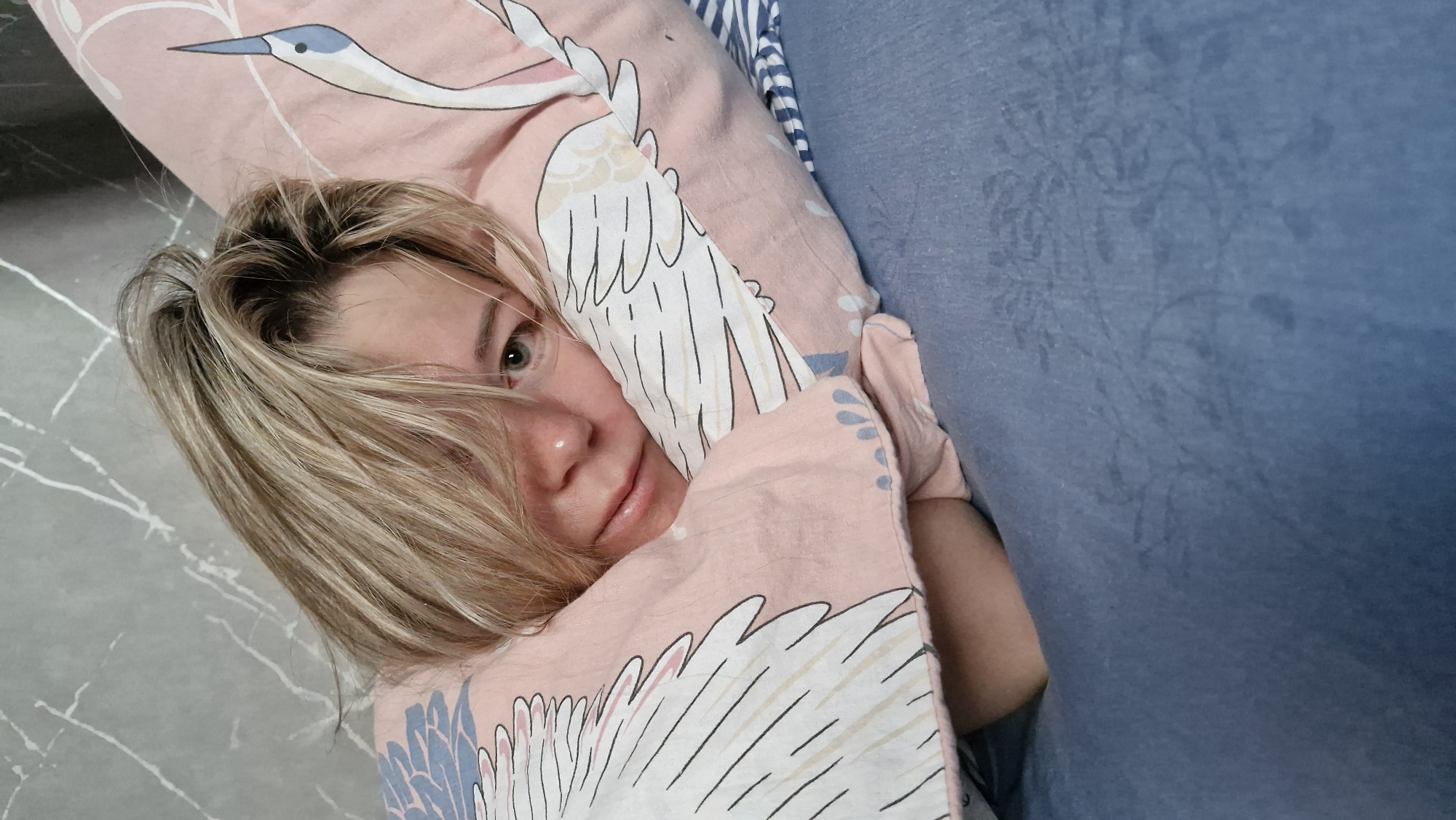 A young woman with fair skin and blonde hair partially covers herself with a patterned pink and white pillow and blanket. The bedding features images of cranes. She is lying on a grey floor with white marbling, next to a blue upholstered object. Her face is visible, and she is looking directly at the camera. The scene appears to be indoors. The location, Otchor, Russia, cannot be independently verified based on the visual information provided.