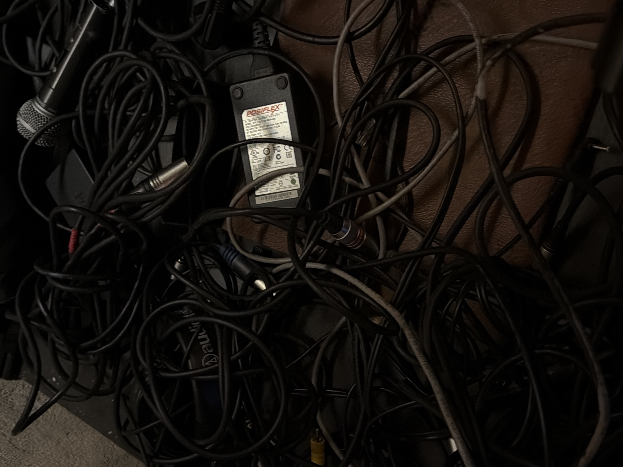 The image presents a close-up, chaotic view of tangled wires and cables. Predominantly black and gray, these cables vary in thickness and type, suggesting a mix of audio, video, and power connections. A microphone is visible on the left side of the image. A black Posiflex power adapter is visible, amidst the tangled cables. The scene appears to be an unorganized storage area, lacking natural light. The precise location within Yerevan, Armenia, is indiscernible from the visual information available.