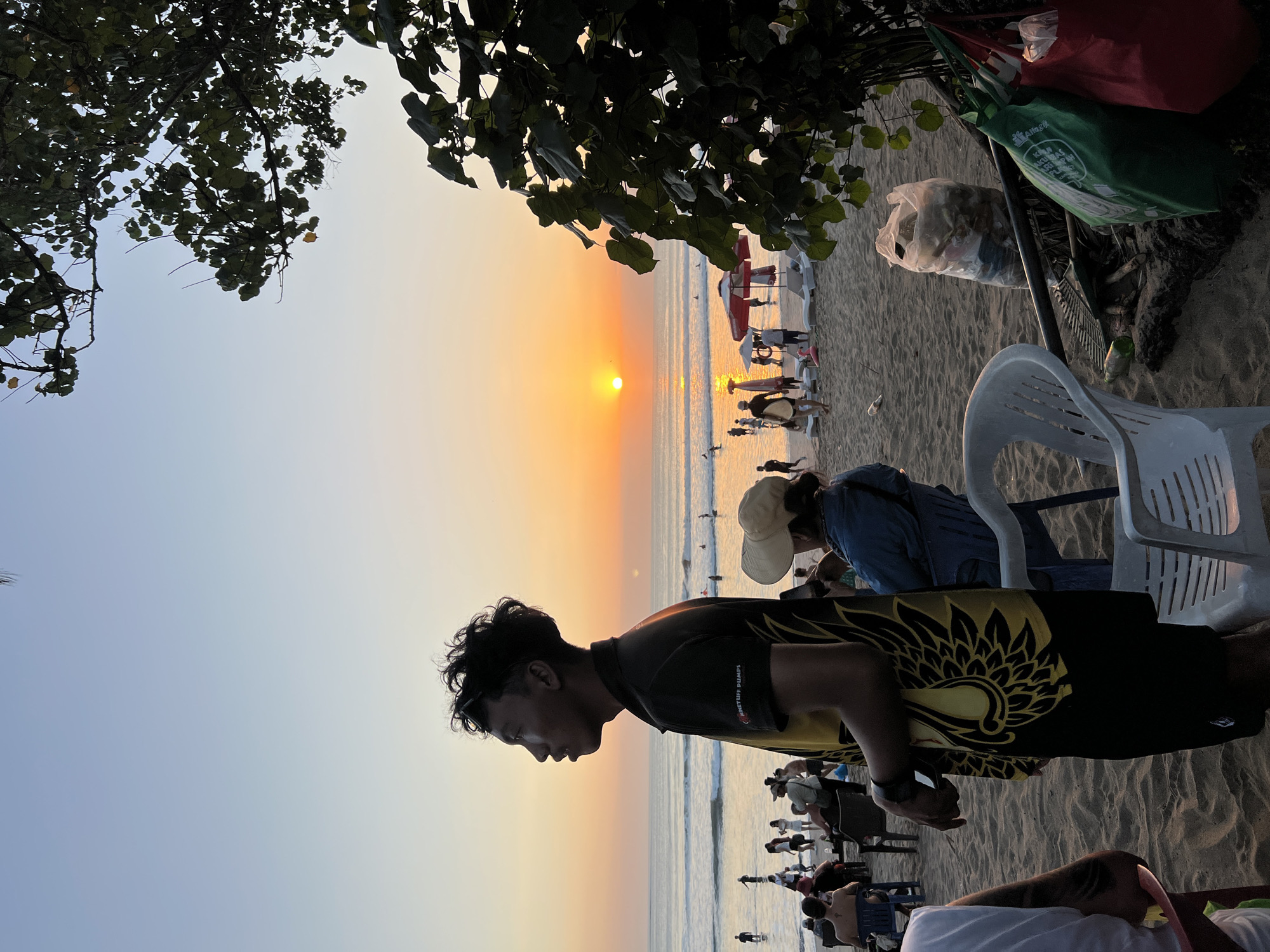 The image captures a beach scene in Kuta, Indonesia, during sunset. In the foreground, a man is standing and looking out towards the ocean, his profile silhouetted against the warm sky. He is wearing a black shirt with a yellow, stylized bird or feather design on the chest and black shorts. Behind him, other beachgoers are seated on chairs or walking along the shore. Some beach umbrellas and lounge chairs are visible. The ocean extends to the horizon, where the sun is setting, casting a golden glow on the water. Trees with large leaves partially frame the right side of the image, and a pile of trash bags is visible on the sand near the foreground. The overall atmosphere is one of a relaxed evening on a popular tourist beach.