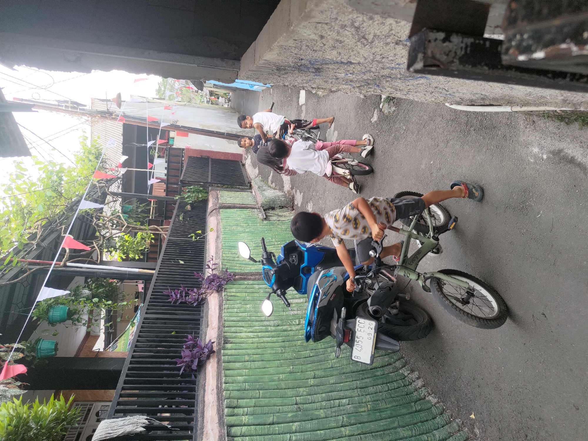 In Depok, Indonesia, on a narrow, paved alleyway, children are seen playing. A young boy, wearing a patterned t-shirt and shorts, is riding a green bicycle with training wheels. To his right, another child, a girl, is riding a pink bicycle, and a boy is on a bike behind her. Further down the alley, more children are visible, with a dark blue motorcycle parked to the left. The alley is lined with walls, one of which has a green, textured surface resembling bamboo. Banners or flags, possibly red and white, are strung across the alley and above the buildings. The environment appears to be a residential area.