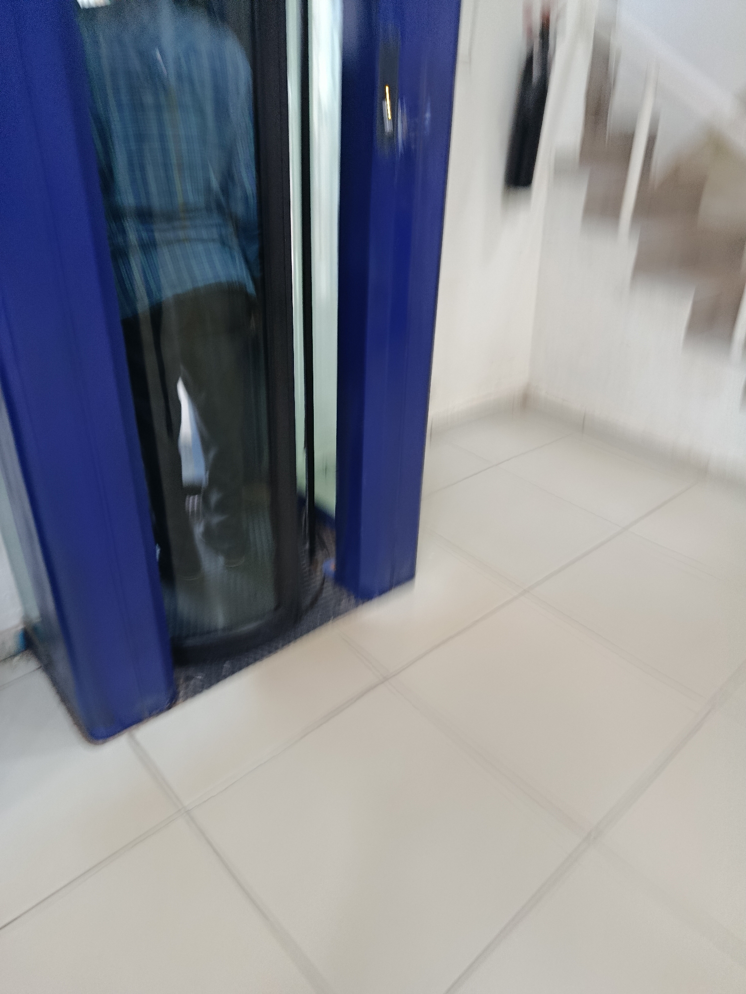 The image shows a blurred view of an interior space. In the foreground, a blue structure, possibly a revolving door or an elevator, occupies the left side of the frame. Inside this structure, a person is visible, wearing a blue plaid shirt and dark pants. The person appears to be in motion, indicated by the blurriness. The floor is tiled with light-colored square tiles. In the background on the right, a staircase with light-colored treads and risers is partially visible, along with a white wall. A dark object, possibly a coat or bag, hangs on the wall near the staircase. The overall impression is of an indoor environment that is either dimly lit or experiencing motion blur. The specific location of Jos, Nigeria, cannot be determined from the visual information present in the image.