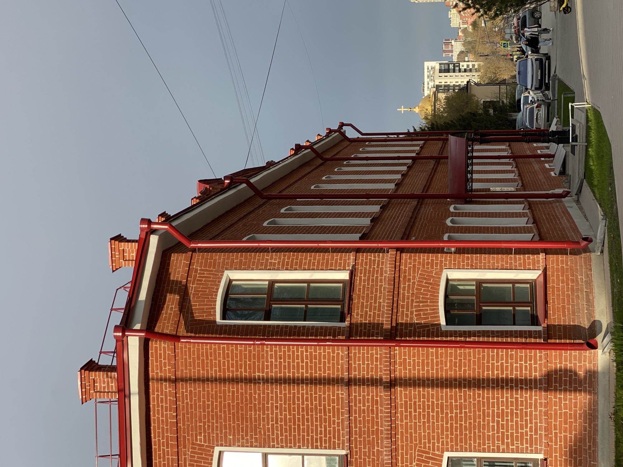 The image presents a side view of a well-maintained, multi-story building constructed from red brick, featuring distinctive red downspouts, pipes, and roof trim. White frames accentuate the rectangular windows on its facade. The reddish-brown roof includes a series of steps or ladders, likely for maintenance, and decorative elements resembling small chimneys on the left side.
The sky is clear and bright, and long shadows are cast across the brick wall, suggesting either early morning or late afternoon sunlight. Several thin power lines traverse the upper portion of the frame.
Adjacent to the building is a paved sidewalk, beyond which lies a street with several parked cars and a few pedestrians in the distance. A yellow electric scooter is partially visible on the far right. In the background, beyond trees and other structures, a modern building rises next to a golden-domed church topped with an Orthodox cross. A visible license plate on a white van in the street, "M205PX 196", confirms the regional code '196', which is associated with Sverdlovsk Oblast, placing the scene in Yekaterinburg, Russia. The image captures a serene urban setting during daylight hours.