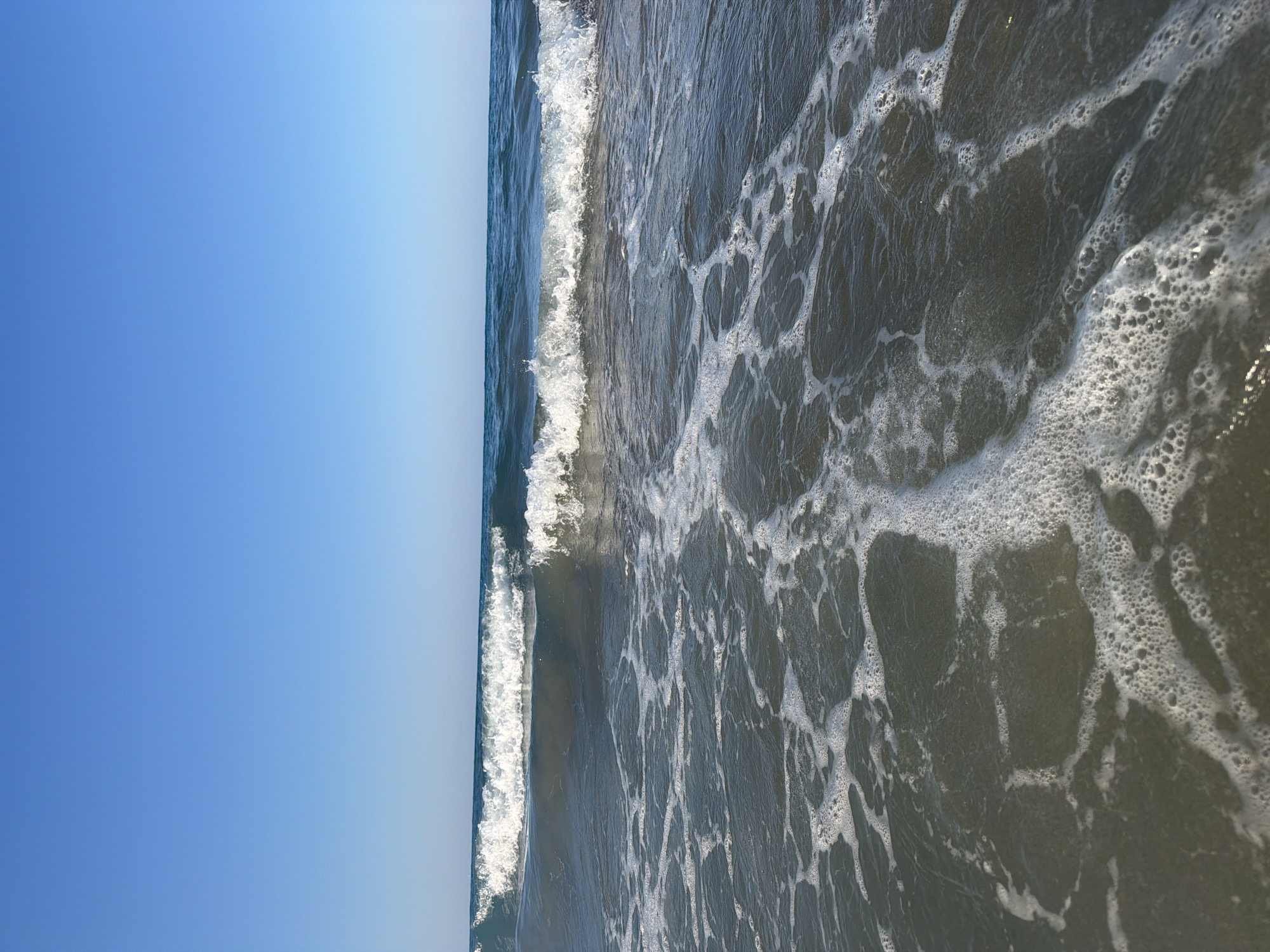 This is a low-angle, eye-level, outdoor, full shot of the sea at Georgioupoli, Greece. The sky is a clear, bright blue with no clouds visible. The horizon line is sharp and straight, dividing the sky from the sea. The sea is a deep blue-green color, with a series of small to medium-sized waves rolling towards the viewer. The waves are breaking, creating white foam and bubbles that are spreading across the surface of the water. The foreground is dominated by the frothy, bubbly water closest to the viewer, with some areas appearing almost white with foam. The lighting suggests it is daytime, likely mid-morning or late afternoon, with the sun casting a slight shimmer on the water's surface. No people or man-made objects are visible in the frame.