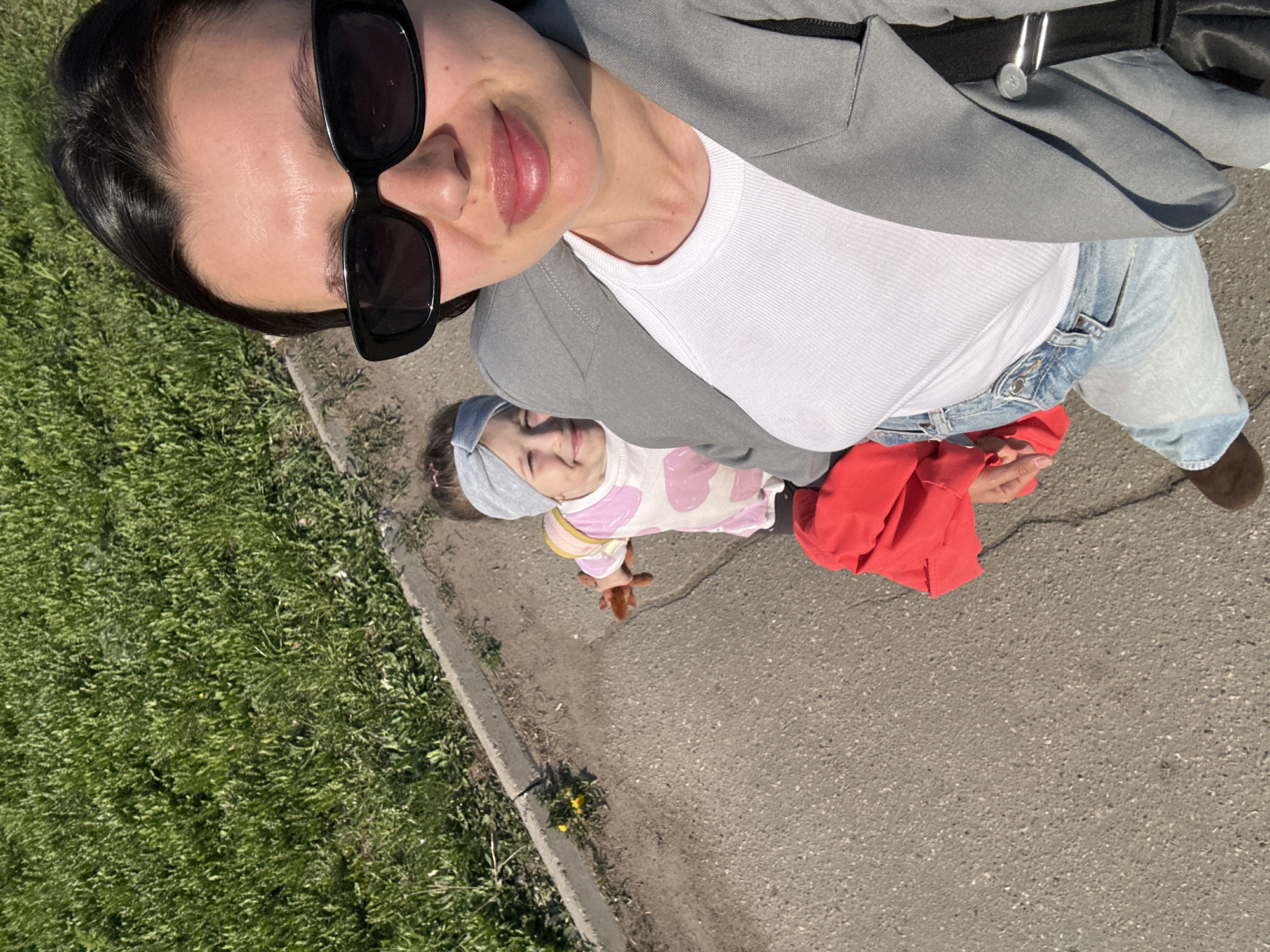 A high-angle photograph depicts a woman and a young child outdoors.
The woman is wearing sunglasses, a white shirt, and a gray jacket. She is also wearing light-colored jeans and brown shoes.
The child is wearing a light-colored outfit with pink hearts on it, and a gray headband. The child is holding a small brown object.
They are standing on a paved surface next to a patch of grass. The location is Chaikovsky, Russia.