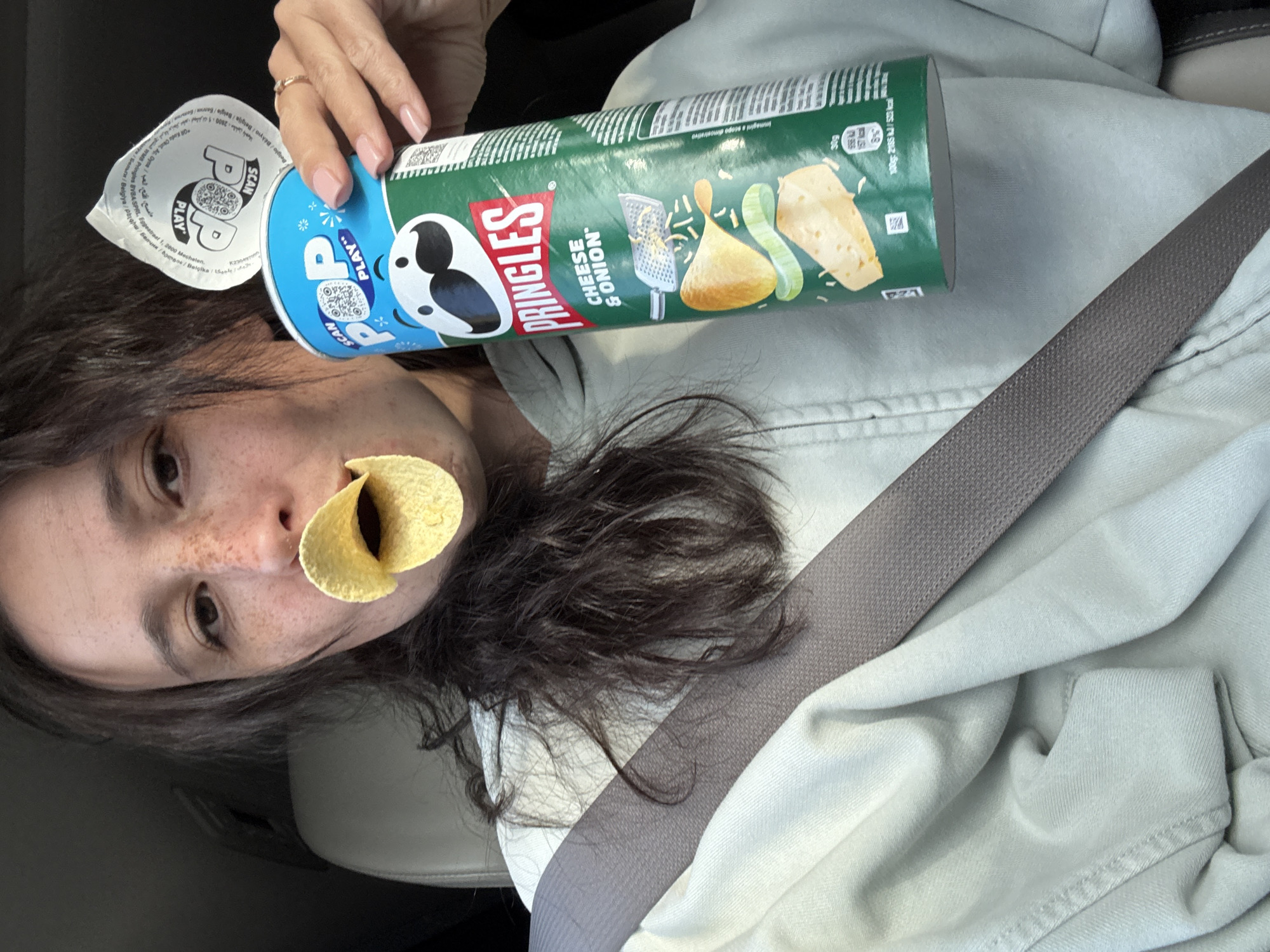 A close-up, tilted shot captures a person inside what appears to be a car, engaged in a playful moment with Pringles. The individual has dark, wavy hair and visible freckles on their nose, with a single Pringles chip playfully positioned in their mouth. Their right hand, adorned with a ring and manicured nails, holds an open green Pringles can.
The can clearly displays the "PRINGLES" logo and the flavor "CHEESE & ONION," along with illustrations of cheese and onions. The blue, peeled-back lid features a "POP PLAY" logo and a "SCAN" prompt with a QR code. Visible text on the can includes nutritional information (e.g., "100g: 2185 kJ/523 kcal") and manufacturer details such as "Pringles BVBA SPRL, Eggestraat 1, 2000 Mechelen," along with multilingual indications of origin including "Belgia / Бельгия."
A grey seatbelt diagonally crosses the person's torso and light-colored clothing, confirming the setting as a vehicle. The background is indistinct, showing parts of the car's interior. The overall scene suggests a casual and lighthearted snack break.