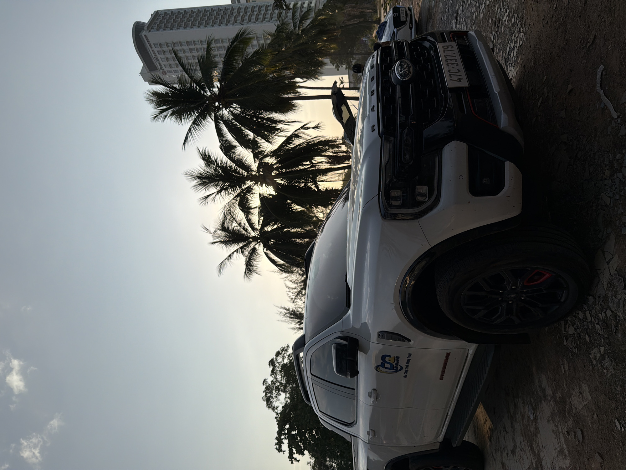 A white Ford Ranger pickup truck is parked on a dirt and gravel lot in Nha Trang, Vietnam. The truck is positioned in the foreground, angled towards the viewer, with its front end prominently displayed. The truck has a black grille with the Ford logo in the center, and the license plate reads "47C-337.79". The truck has a "Stormtrak" decal on the lower door. In the background, palm trees sway in the breeze, and a tall, modern hotel with a curved top rises into the sky. The sky is a pale blue with a few wispy clouds, illuminated by the setting sun which casts a warm glow. Another car, a white Mazda, is parked to the right of the Ford Ranger. The overall scene suggests a tropical coastal setting, likely during a late afternoon or early evening.