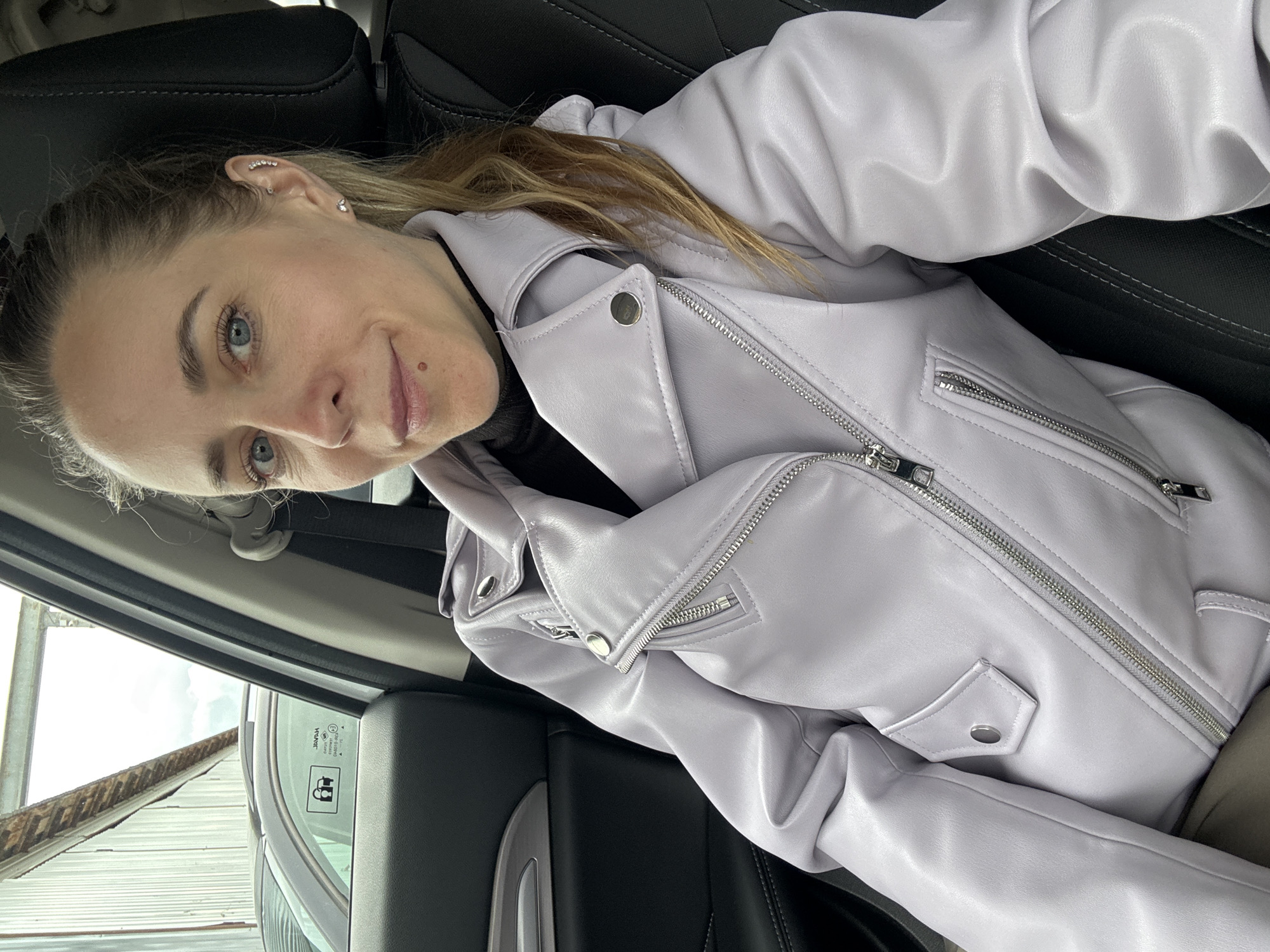 A woman with blonde hair pulled back is seated in a car, wearing a light purple leather jacket. She is looking at the camera with a slight smile. The background shows the interior of the car and a glimpse outside the window, revealing a building with a corrugated metal roof. Although the image does not definitively confirm the location, the request includes the city of Chaikovsky, Russia. Therefore, it can be assumed that the photo was taken in or near Chaikovsky.
