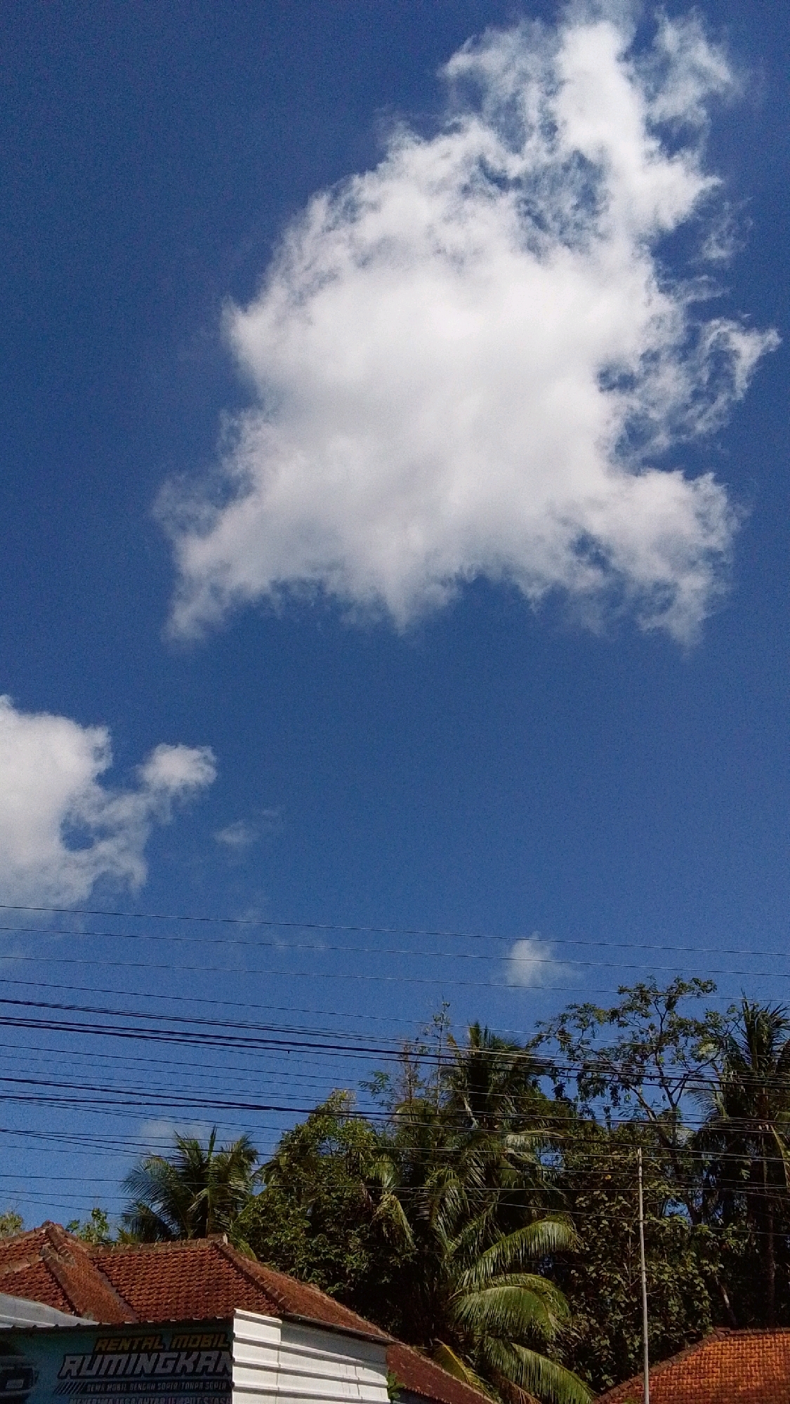The image depicts a clear blue sky with scattered white clouds. In the foreground, there is a brown tiled roof of a building, likely a house or a shop. Below the roof, a portion of a white facade is visible, with some lettering that appears to read "RENTAL MOBIL" and "RUMINGKAN". Power lines are strung horizontally across the mid-section of the image, against the backdrop of the sky and lush green trees that rise behind the building. The foliage suggests a tropical or subtropical environment, consistent with locations like Cintaratu, Indonesia. The scene appears to be an outdoor view, possibly taken during the daytime, given the bright sky and prominent clouds. There are no people or animals visible in the frame. The overall impression is of a serene, everyday urban or semi-urban landscape.