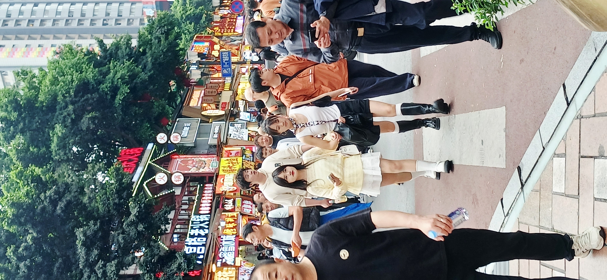 The image captures a vibrant street scene in Yuzhong District, China. A group of pedestrians of varying ages are walking or standing along a pedestrian brick-paved street. In the background are buildings adorned with bright signage and neon lights, with a portion of the structures hidden by lush green trees. The environment suggests a commercial or tourist area.