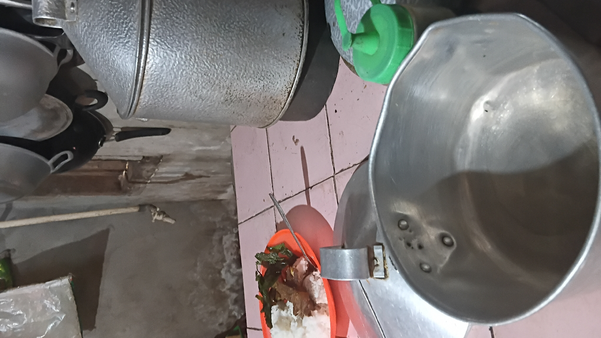 The image depicts a kitchen scene, possibly in Prabumulih, Indonesia, featuring several cooking utensils. In the foreground, a large, unwashed aluminum pot is positioned prominently, its interior reflecting the ambient light. To the left, a silver lid rests on a surface. Further back, an orange plate holds a portion of rice with meat and leafy green vegetables, accompanied by a silver spoon. Dominating the right side of the image is a large, textured aluminum pot with a lid, situated on a dark base. Behind this, a green plastic bottle with a nozzle is visible. The background shows a wall with a faucet and pipes, and several hanging pots and pans. The overall environment appears to be functional and utilitarian, typical of a kitchen space.