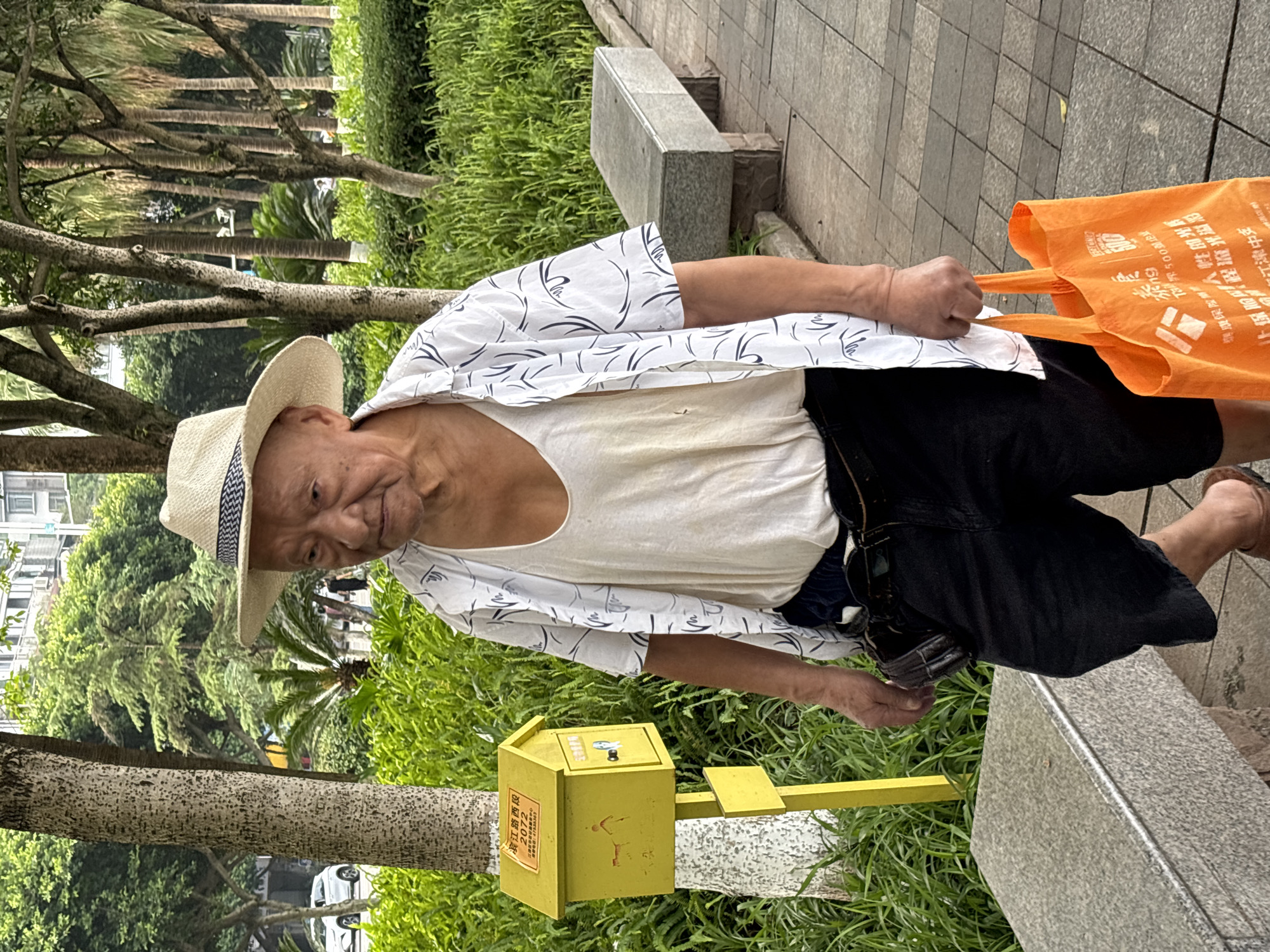 An elderly East Asian man wearing a straw hat, a white tank top, a white short-sleeved shirt with a black pattern, and black shorts walks on a paved sidewalk in a park-like setting. He is holding an orange reusable shopping bag in his right hand. To his left, there is a yellow mailbox on a pole, and behind him, there are trees and a stone bench. The scene appears to be in the city of Jiangjin, China, as indicated by Chinese text on the mailbox and the shopping bag. The man has a serious expression on his face.