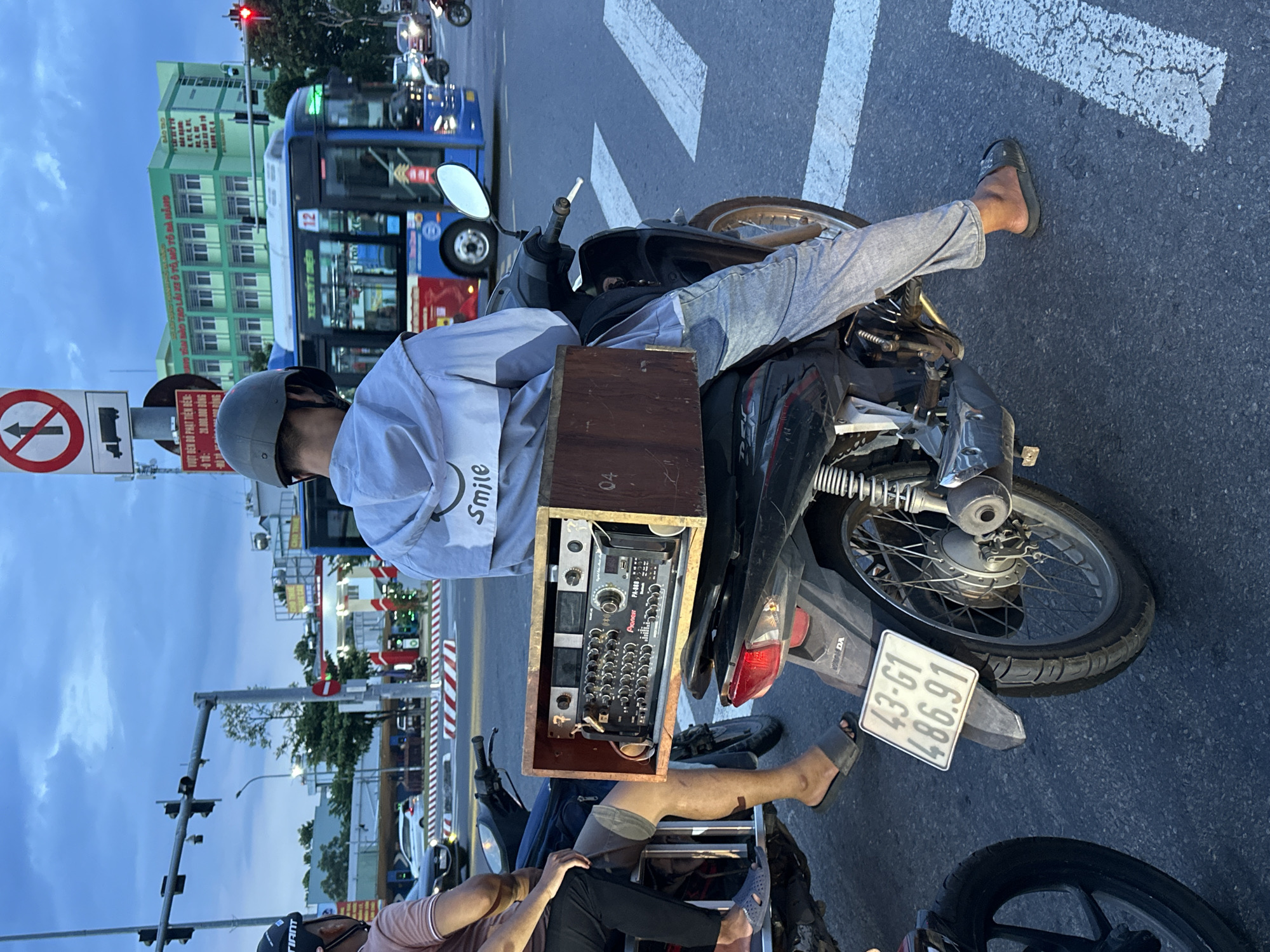 This image captures a street scene in Đà Nẵng, Vietnam, likely at an intersection. In the foreground, two individuals are seated on motorcycles. The person on the right, wearing a light-colored hooded jacket with a "Smile" graphic and a gray helmet, is the primary subject. Their motorcycle has a wooden box mounted on the back, containing electronic audio equipment, including a Pioneer PA-600 amplifier. The license plate of this motorcycle reads "43-G1 486.91." The person on the left, partially visible, is wearing a dark helmet and a pink shirt. The background shows a bustling city environment with traffic lights, a blue bus with the number "12" displayed, other vehicles, and a multi-story green building with Vietnamese signage. The sky is overcast, suggesting it might be early morning or late afternoon. The scene is set on a paved road with pedestrian crossing markings.