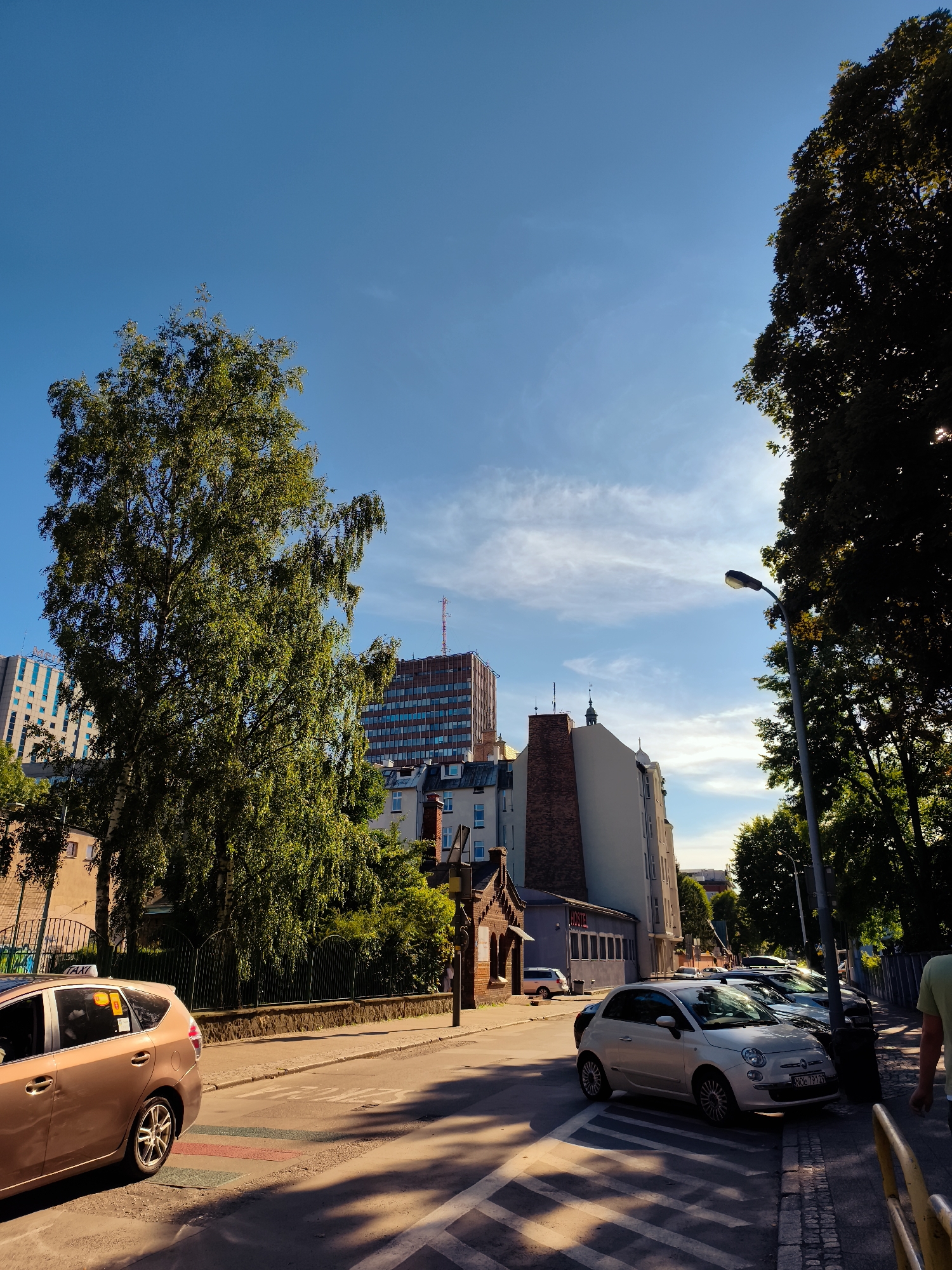 This is a photograph taken on a sunny day in the city of Gdańsk, Poland, depicting a street scene. The perspective is from the sidewalk, looking down a street lined with trees and buildings. In the foreground, a beige taxi is parked on the left, partially obscuring a crosswalk. Further down the street, several cars are parked along the curb, including a white Fiat 500. The street is paved, with a sidewalk made of cobblestones and concrete.
The buildings on the right side of the street are a mix of architectural styles, with some older brick structures and taller, more modern buildings in the background. One notable building in the background appears to be an office building or hotel with a repeating pattern of windows. A large tree with green foliage dominates the left side of the frame, casting shadows on the street. The sky is a clear blue with a few wispy clouds. A street lamp is visible on the right side of the frame. A person is partially visible on the far right edge of the frame, walking away from the camera. The image conveys a sense of urban life on a pleasant day.