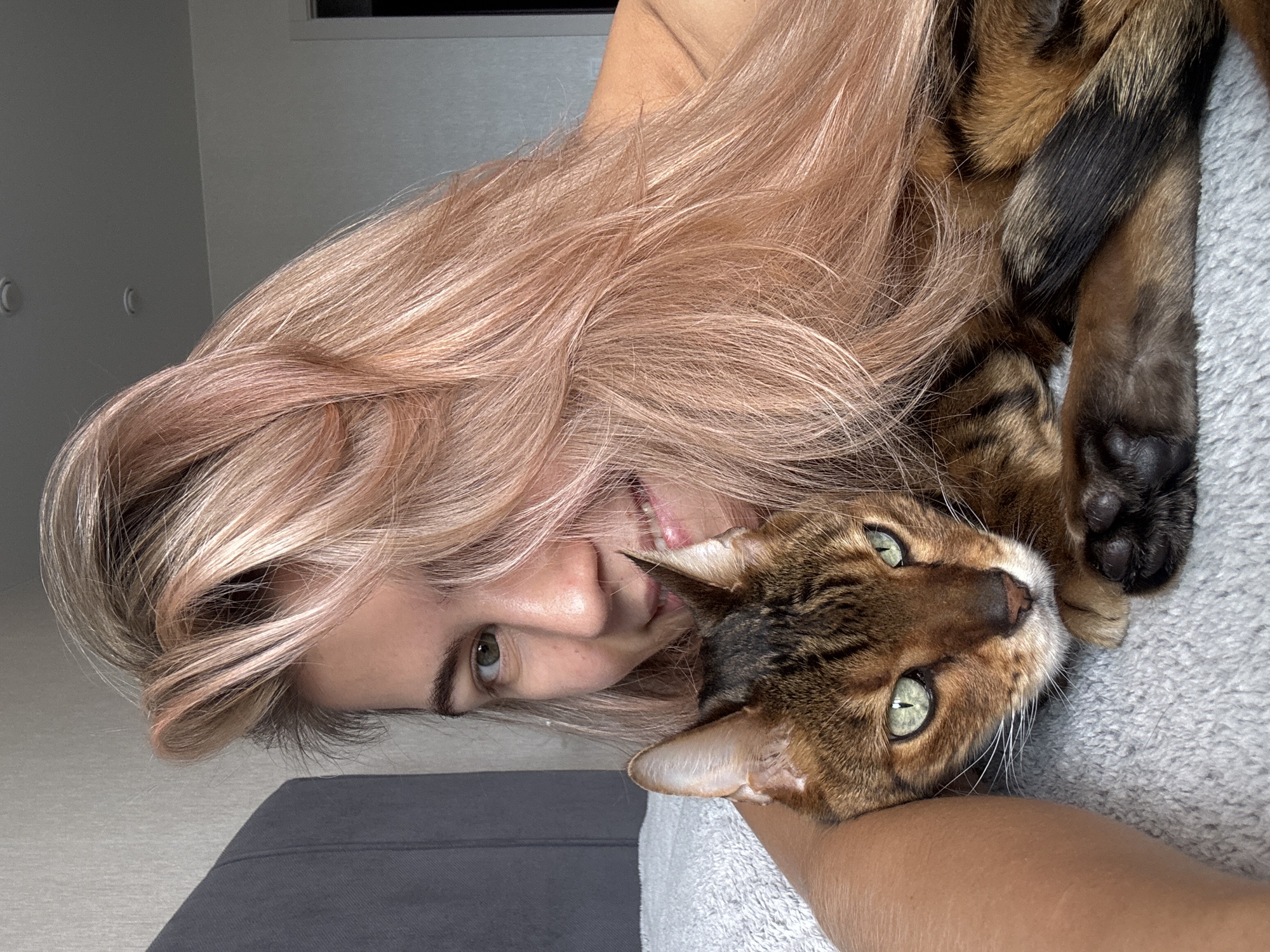 A woman with pinkish-blonde hair is holding a Bengal cat. The woman is smiling slightly and looking at the camera. The cat is lying on its side, with its head resting on the woman's arm. The background is blurry and appears to be an interior. The location is Unknown, Unknown.