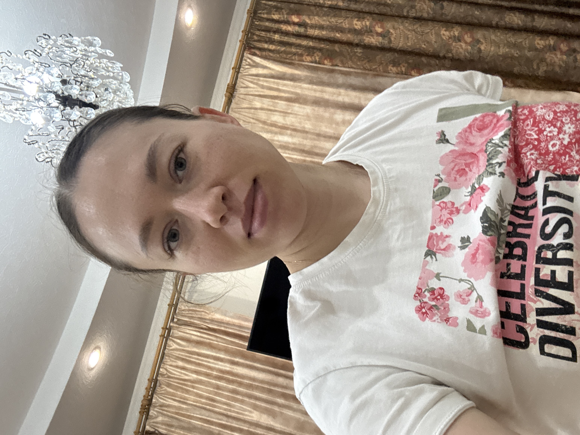 The image shows a person wearing a white t-shirt with a floral design and the words "CELEBRATE DIVERSITY" printed on it. The person has fair skin, brown hair pulled back, and is looking directly at the camera. The background includes a decorative ceiling light fixture and beige, patterned curtains. A dark, flat screen appears behind the person. The location is possibly in Ферма, Russia.