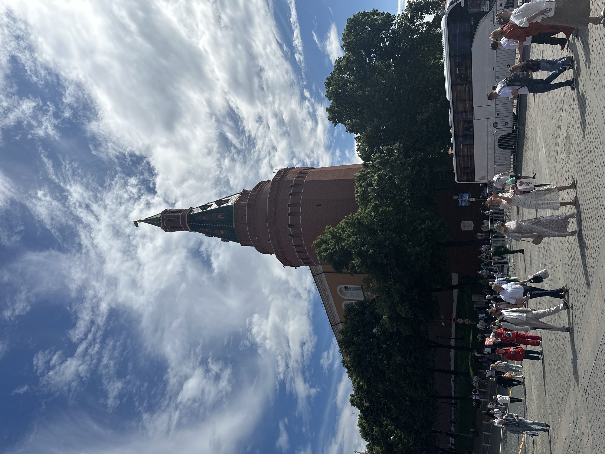 The media file shows a low-angle, vertical shot of a red brick tower with a green roof, likely a historic landmark in Moscow, Russia, under a bright blue sky filled with scattered white clouds. The tower's architectural details, including arched windows and decorative elements, are visible. Lush green trees frame the base of the tower, and a section of a yellow building with arched windows is also visible.
In the foreground and to the right of the tower, a group of people are standing and walking on a paved area. Some individuals are dressed in casual wear, while others are in more formal attire. A white tour bus is parked behind some of the people. The scene appears to be an outdoor tourist area, with the presence of a bus suggesting a tour group. The lighting indicates it is daytime, likely late morning or afternoon, with clear, sunny weather. The overall atmosphere seems to be that of a busy tourist attraction.