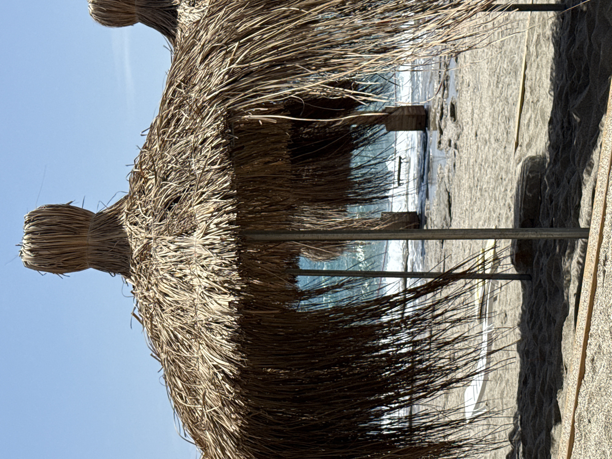 The image captures a beach scene in Alanya, Turkey, viewed from under a thatched beach umbrella. The scene includes a sandy beach, the Mediterranean Sea, and clear blue sky. The umbrella, constructed from dried grasses, provides shade and dominates the foreground. The beach appears lightly populated, with indications of beach furniture present. The overall environment suggests a sunny, coastal resort atmosphere.