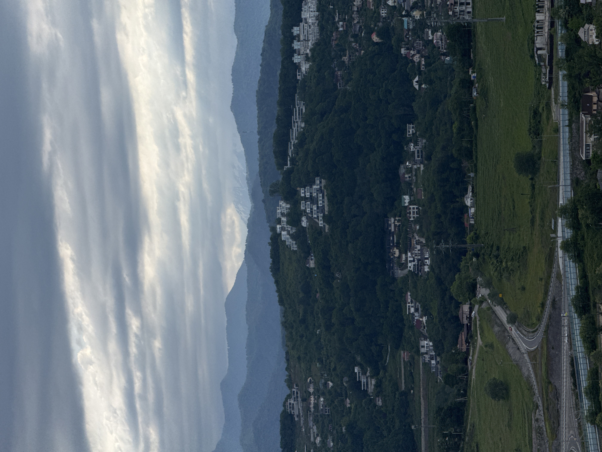 The image presents a landscape featuring the city of Sochi, Russia. Mountains ascend from the left, marked by layers of grey and blue hues, potentially indicating distance and atmospheric perspective. The foreground shifts towards the right, transitioning into dense, dark green vegetation interspersed with white buildings and residences, typical of an urban settlement built on inclined terrain. A section on the right side of the image shows a road with a series of cars on the right-most road, and a grassy area. The sky, visible on the far left, is filled with clouds. The photograph gives a sense of the mountainous terrain of the area.