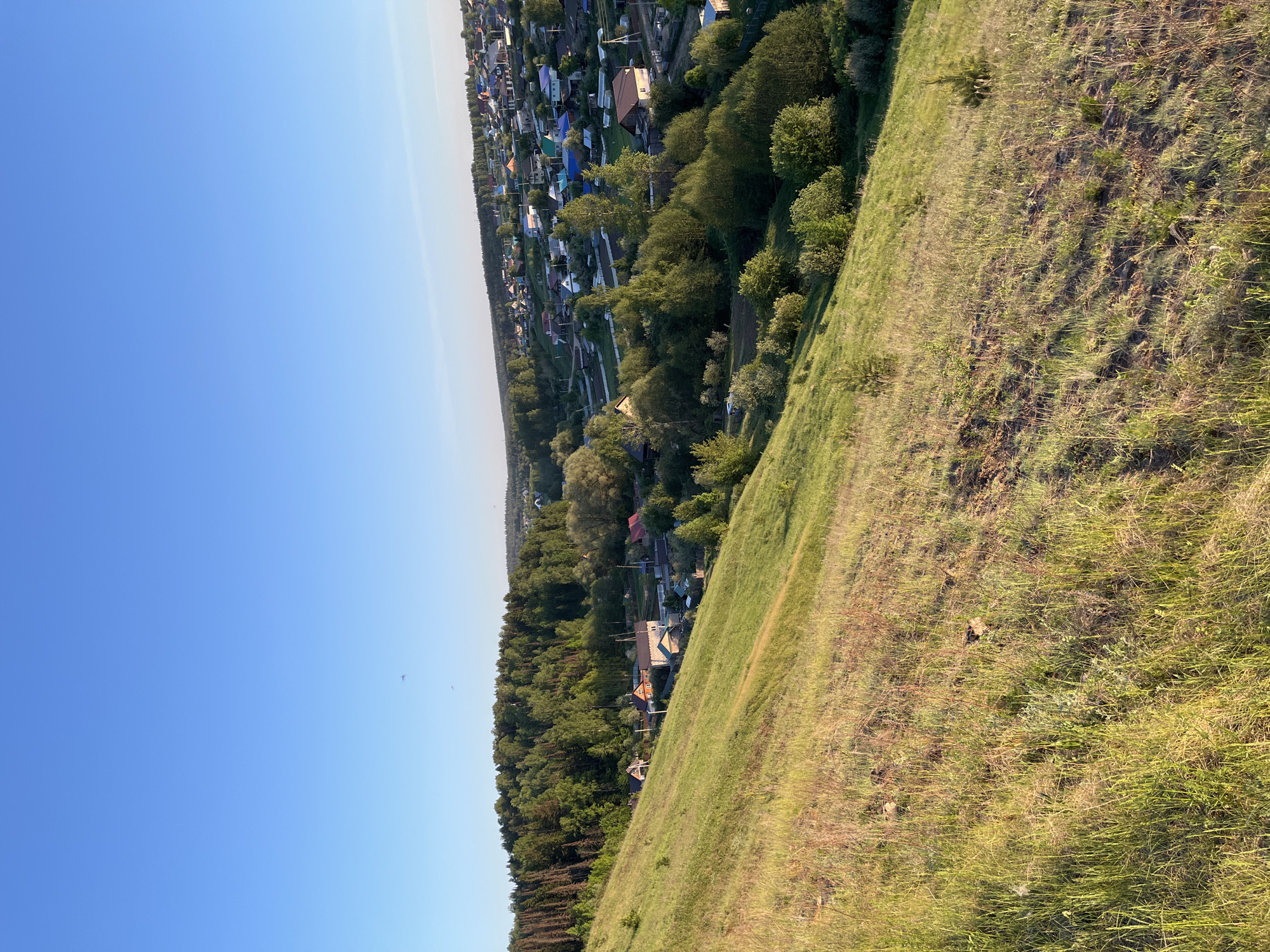 The image shows a rural landscape in Ольховка, Russia, taken from an elevated position on a grassy hillside. The foreground is dominated by the hillside, covered in grasses. Beyond the hillside, a line of trees separates it from a village. The village consists of numerous houses with various colored roofs. The sky is clear and blue.