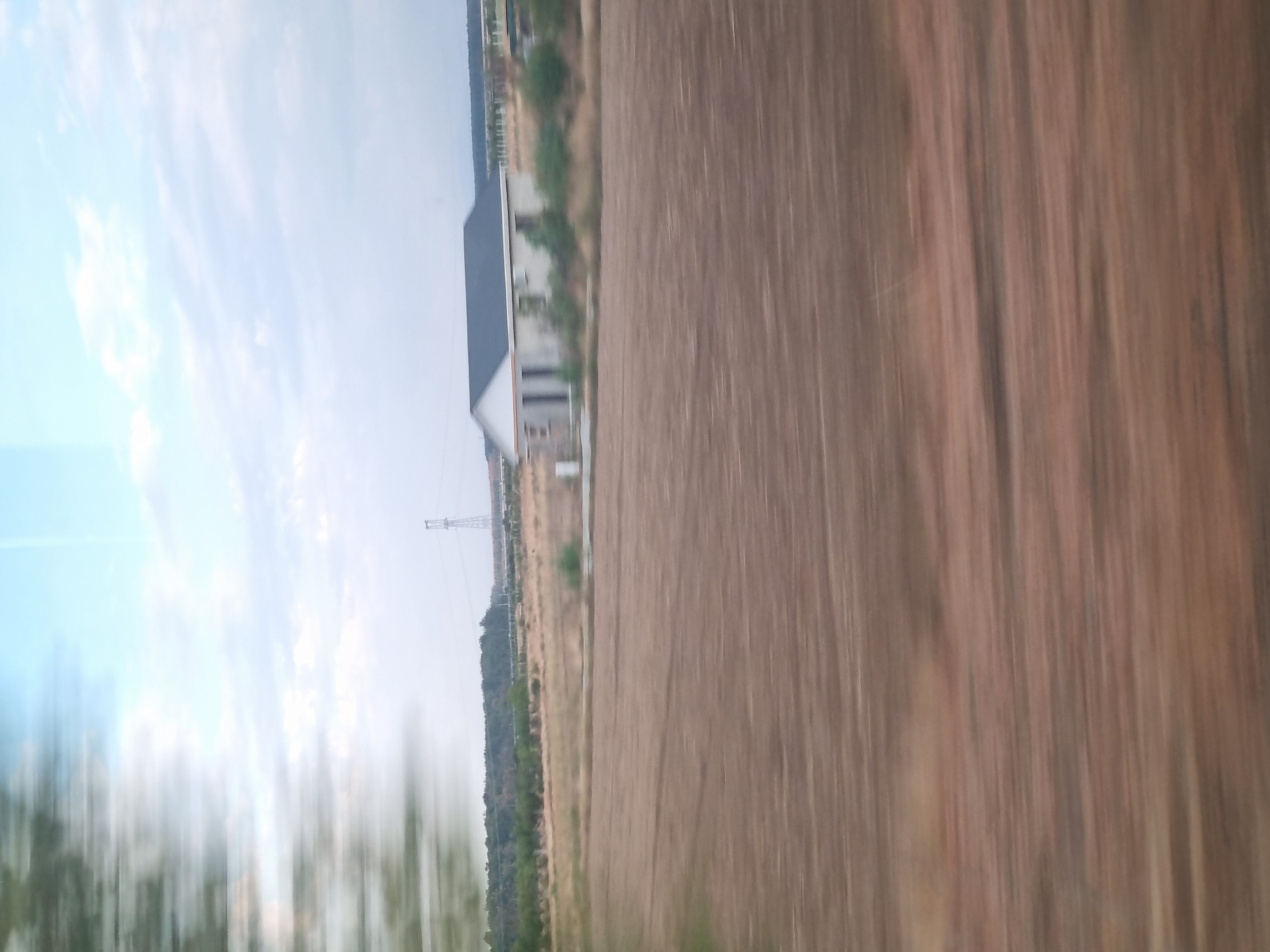 A blurred landscape view from a moving vehicle shows a residential building with a dark roof and white walls, situated in an open, agricultural area. The ground in the foreground appears to be plowed earth, with visible furrows. In the middle ground, dry grass and scattered bushes surround the house. In the background, a line of trees marks the horizon, with a power line tower visible in the distance. The sky is overcast with a pale blue hue. The image was captured in Gepiu, Romania.