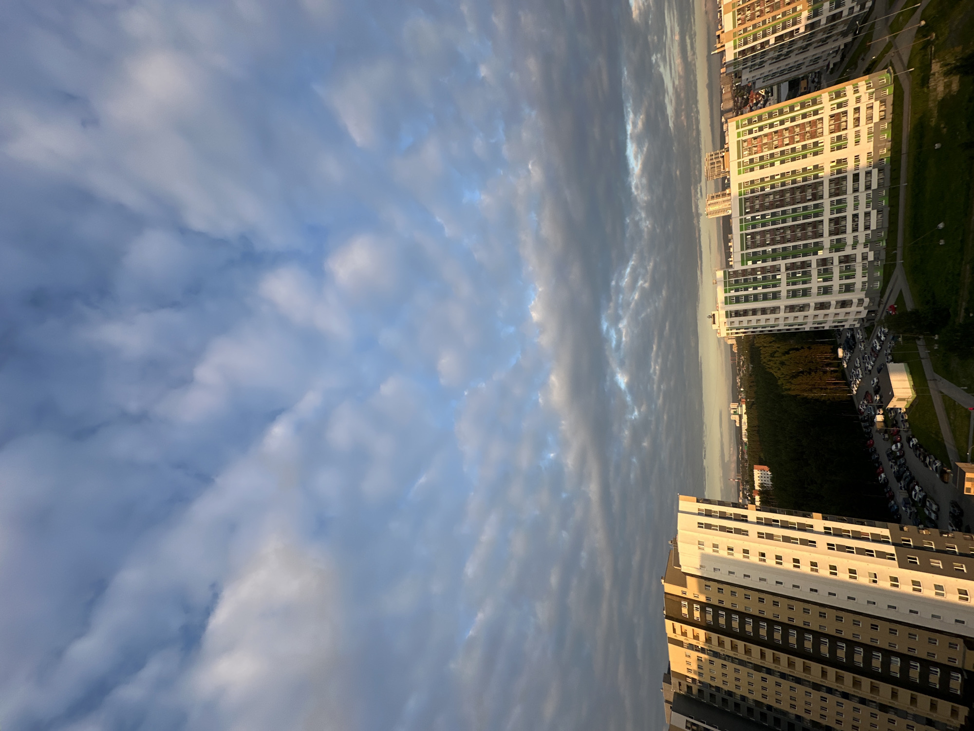 This is an aerial photograph of the southern district of Yekaterinburg, Russia, specifically the "Botaniсhesky" residential complex. The image captures the late afternoon sky filled with scattered clouds, casting shadows on the modern multi-story apartment buildings below. The buildings are characterized by their varied architectural styles and colors, with some featuring prominent green accents. A dense forest separates the residential area from the more distant cityscape, which is visible on the horizon under the softening light. A large parking lot filled with cars is visible adjacent to one of the buildings, indicating a populated urban environment. The overall scene depicts a contemporary residential area within a large Russian city.